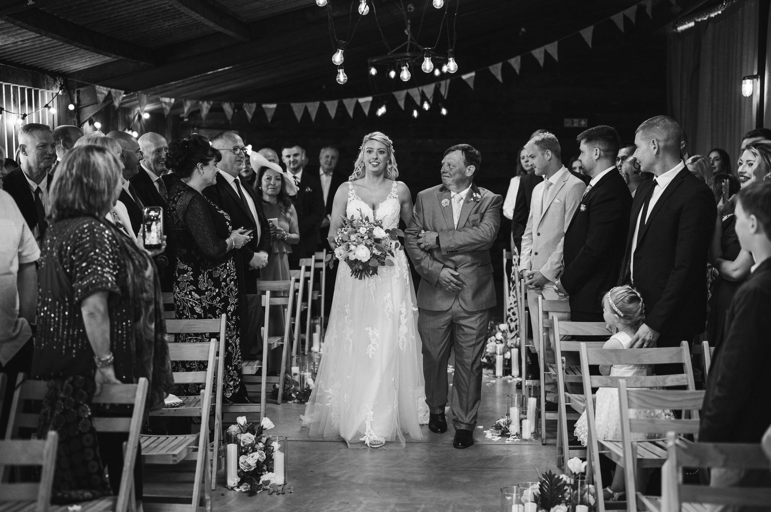 Black and white photo of a wedding ceremony with a bride walking down the aisle accompanied by an older man, possibly her father, in a rustic indoor venue grange barn Whitchurch, Gareth roy photography