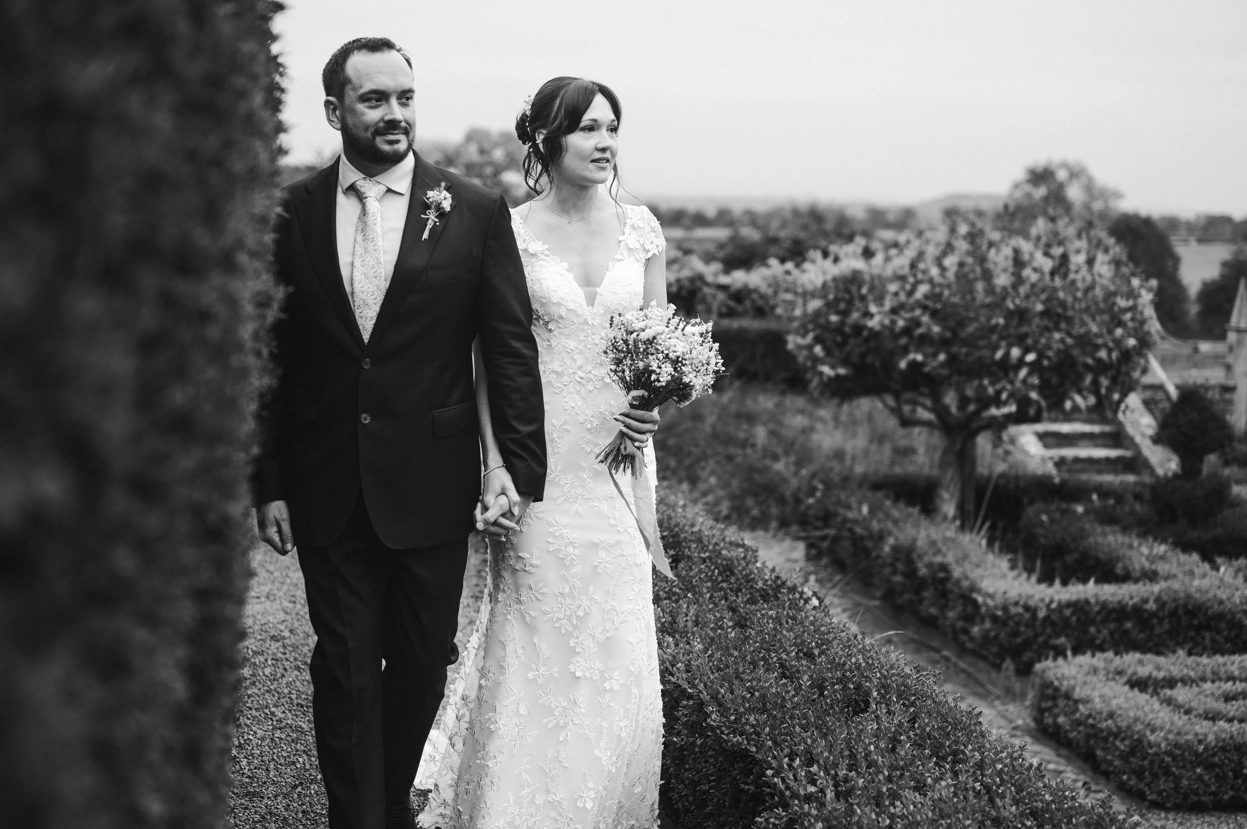 A black-and-white photo of a bride and groom walking hand in hand through a garden, with the groom in a suit and the bride holding a bouquet of flowers pauntley court, Gareth roy photography