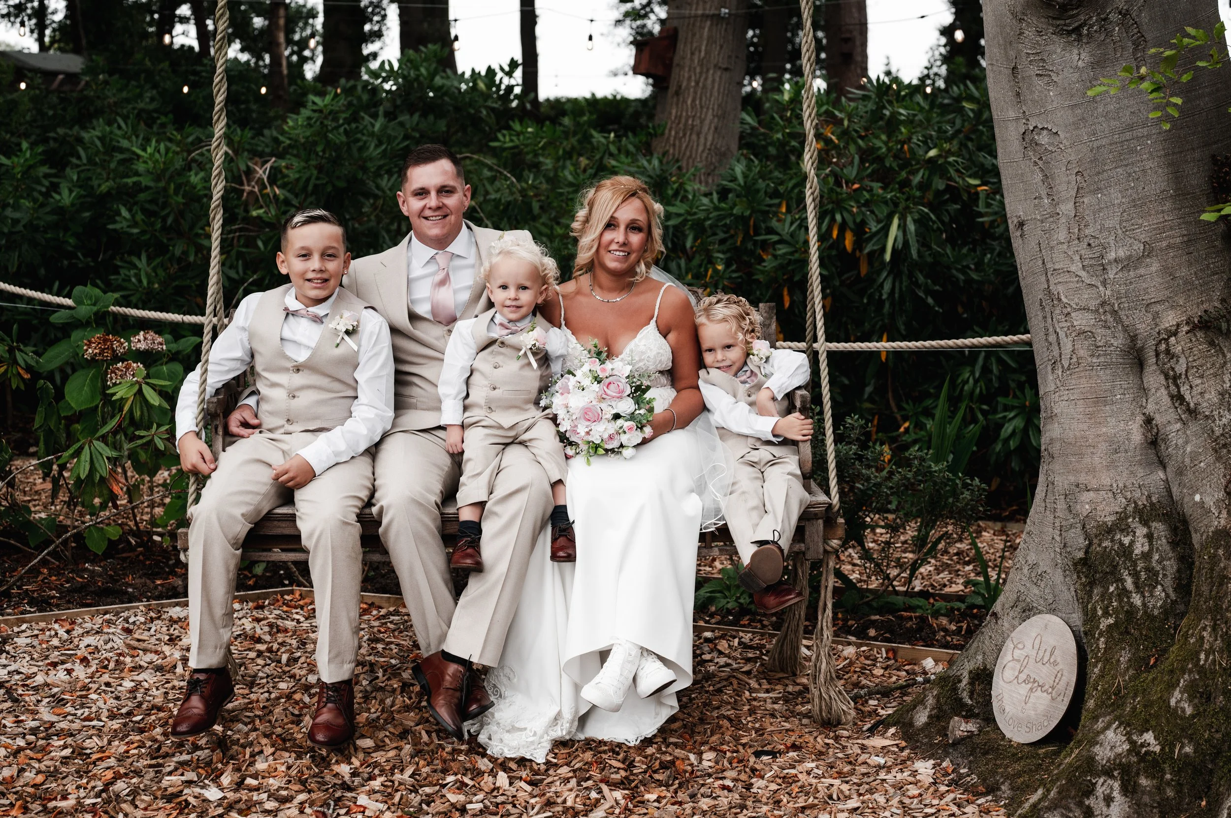 The Love Shack - Groom and bride in wedding attire, sat on a wooden swing with three young children. 