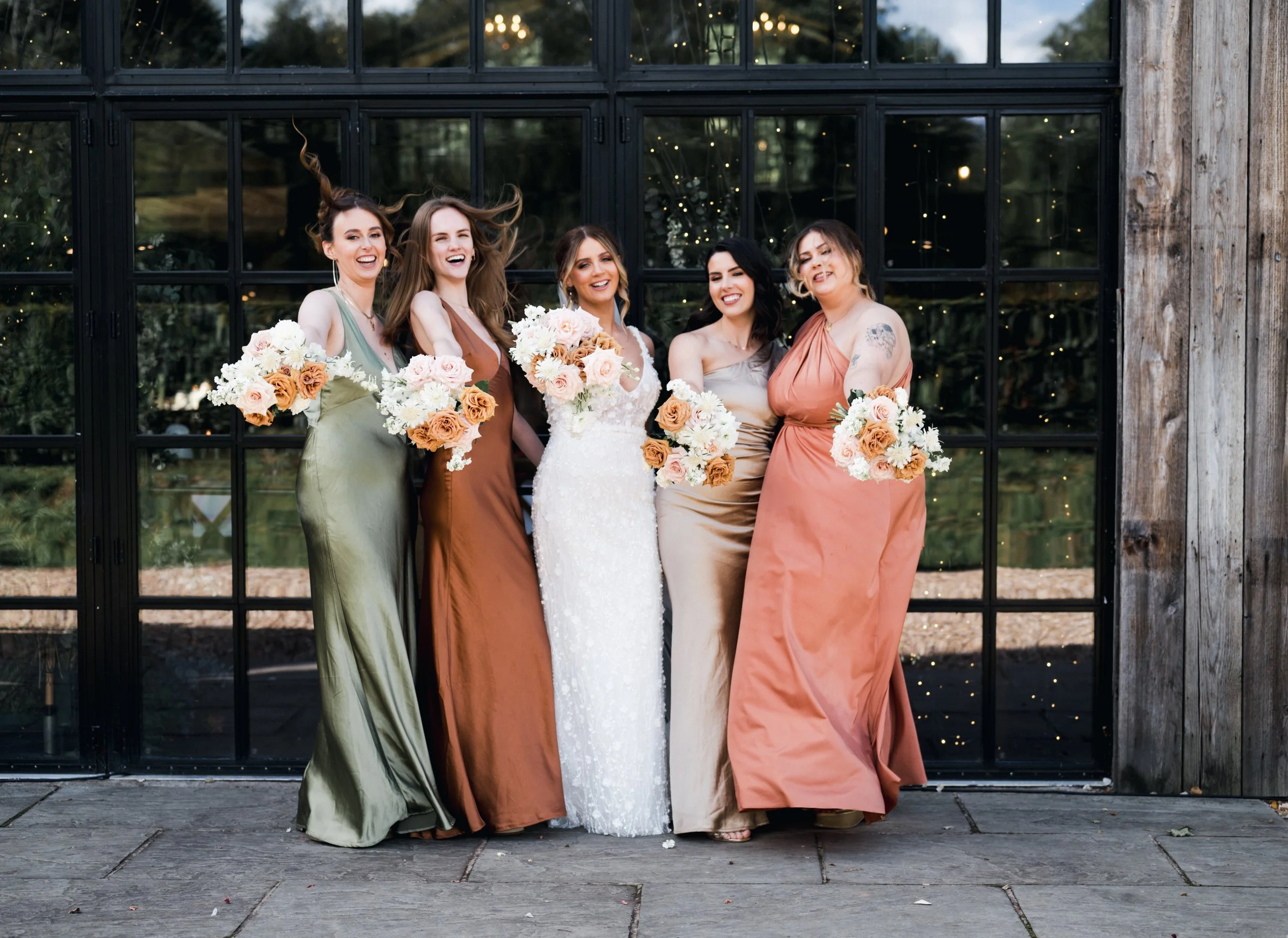 Five women, including a bride in a white wedding dress, standing in front of a black-framed glass door at a wedding reception, holding bouquets of peach and cream flowers, smiling and posing for the photo. hidden river cabins, Gareth roy photography