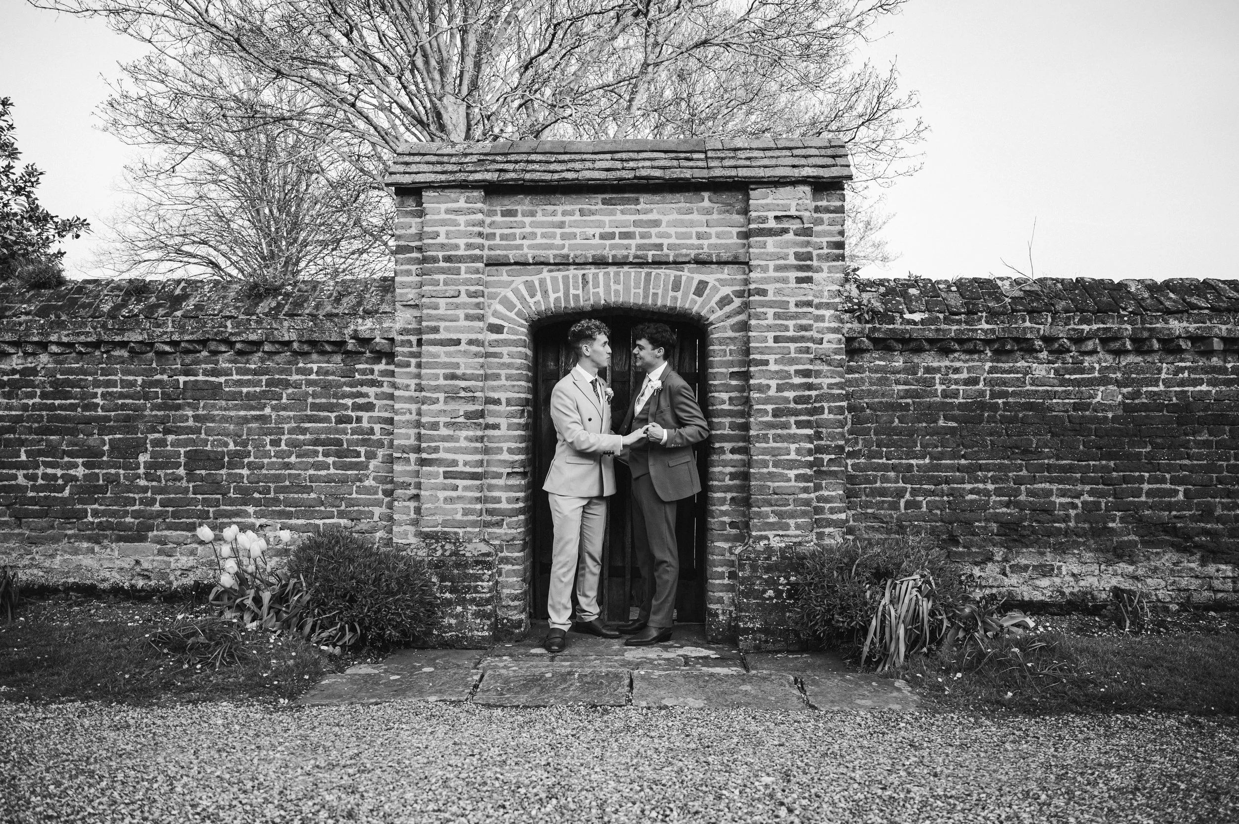 Black and white image of two grooms holding hands in a brick archway