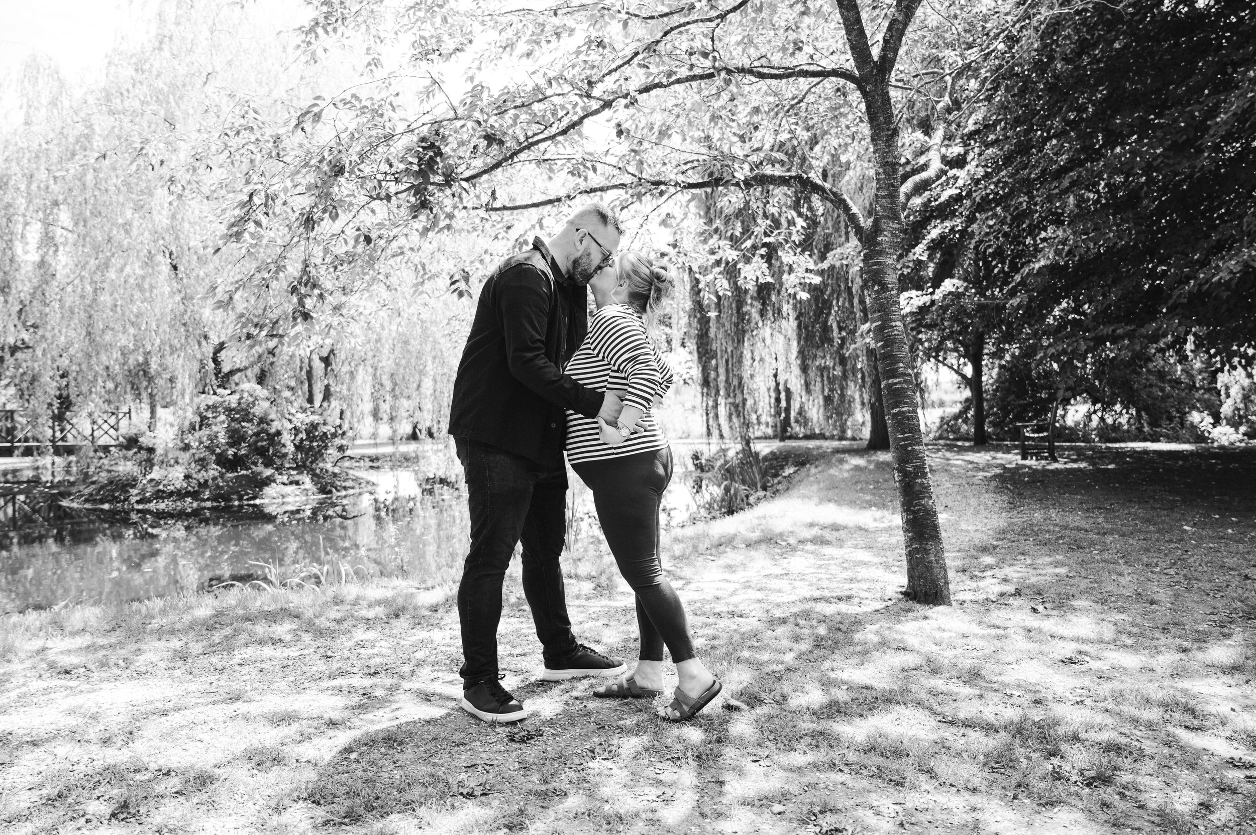 The Grosvenor Pulford Engagement - black and white image of man in black shirt and woman in striped top, embracing kissing, surrounded y greenery and trees with lake behind them. 