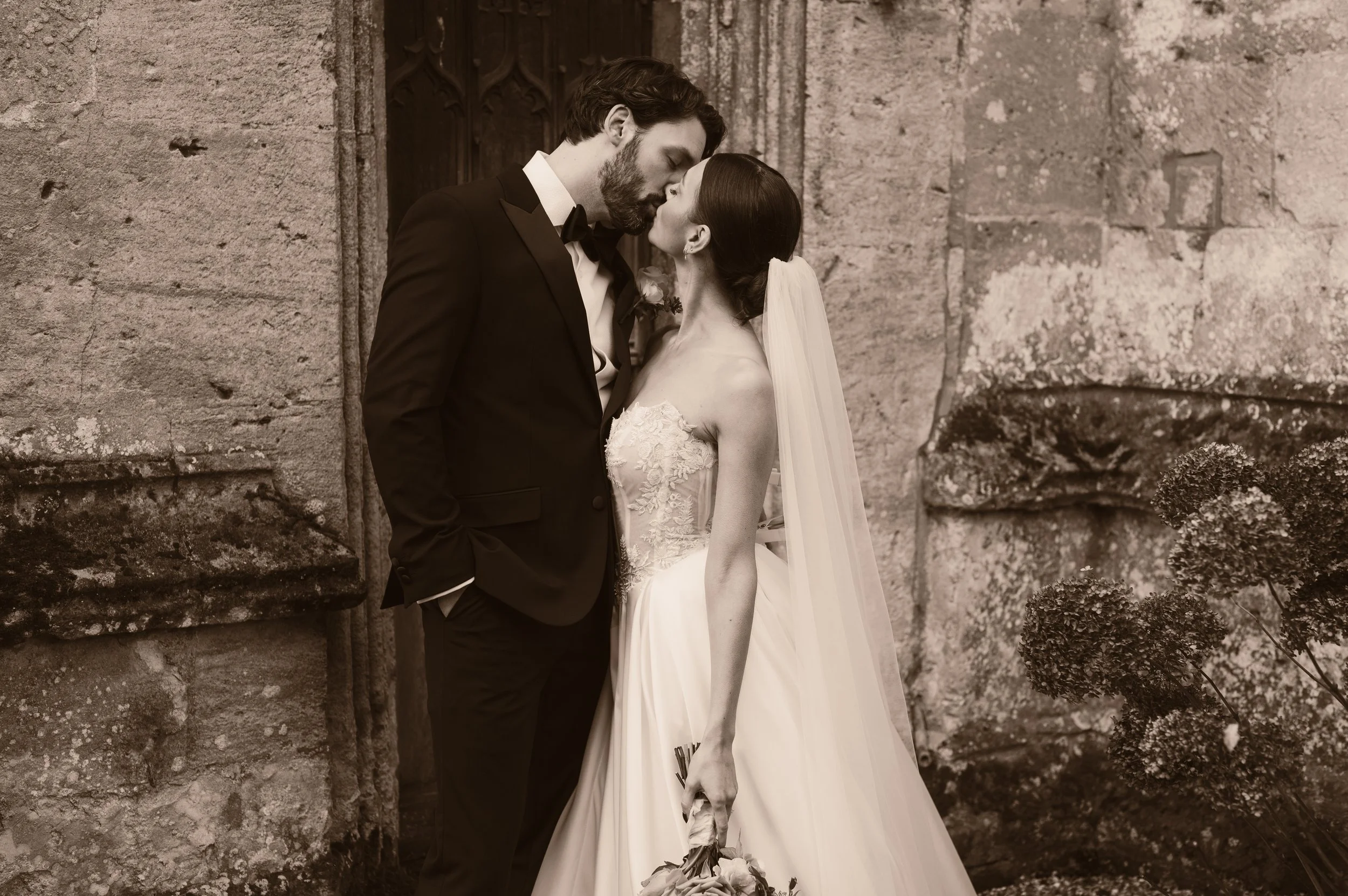 A bride and groom share a kiss outdoors in front of an old stone building, the bride holding a bouquet, both dressed in wedding attire at sudeley castle, Gareth roy photography