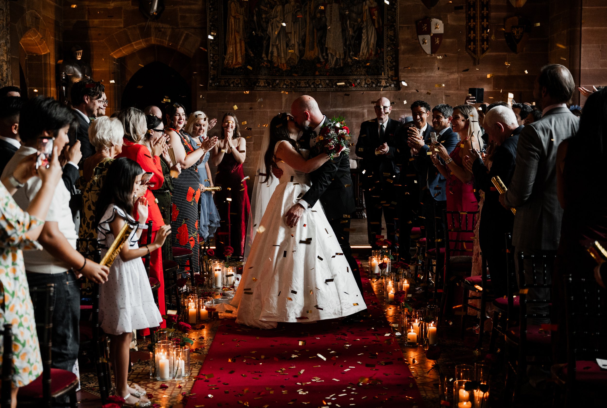 Peckforton Castle - bride and groom in wedding attire kissing after wedding ceremony, surrounded by wedding guests and confetti in the air above them. 