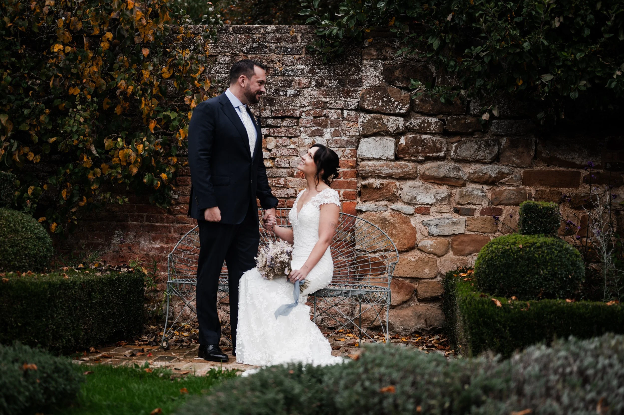 A bride and groom holding hands in a garden. The bride is sitting on a metal bench, holding a bouquet, and looking up at the groom, who is standing and smiling pauntley court, Gareth roy photography