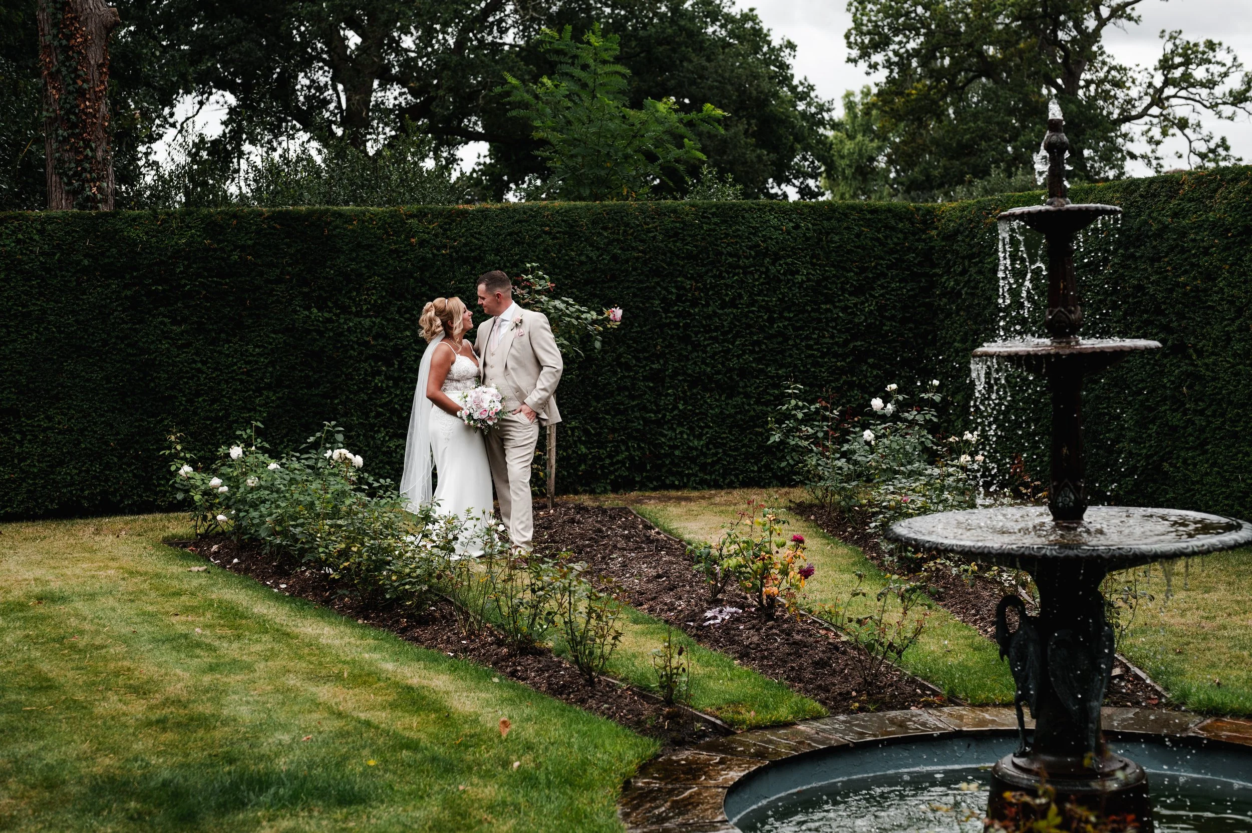 The Love Shack - Groom and bride in wedding attire, walking through gardens towards a fountain.  