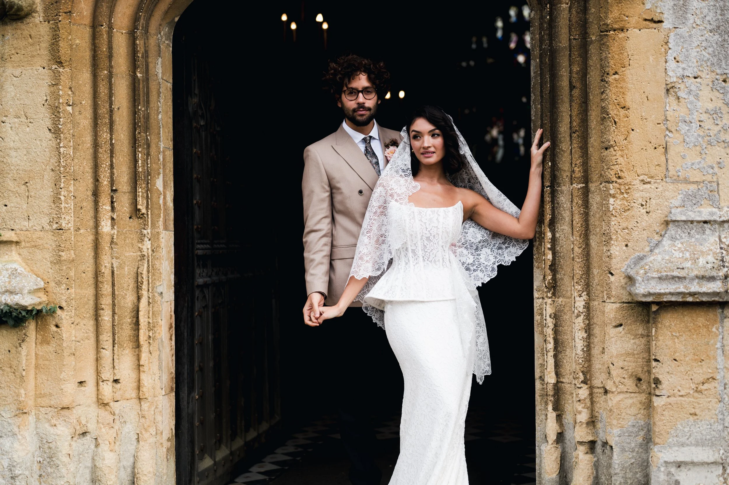A bride and groom standing at a stone archway, holding hands, with the bride wearing a lace wedding dress and veil, and the groom in a beige suit. The background inside the arch is dark, with some blurred lights visible at sudeley castle