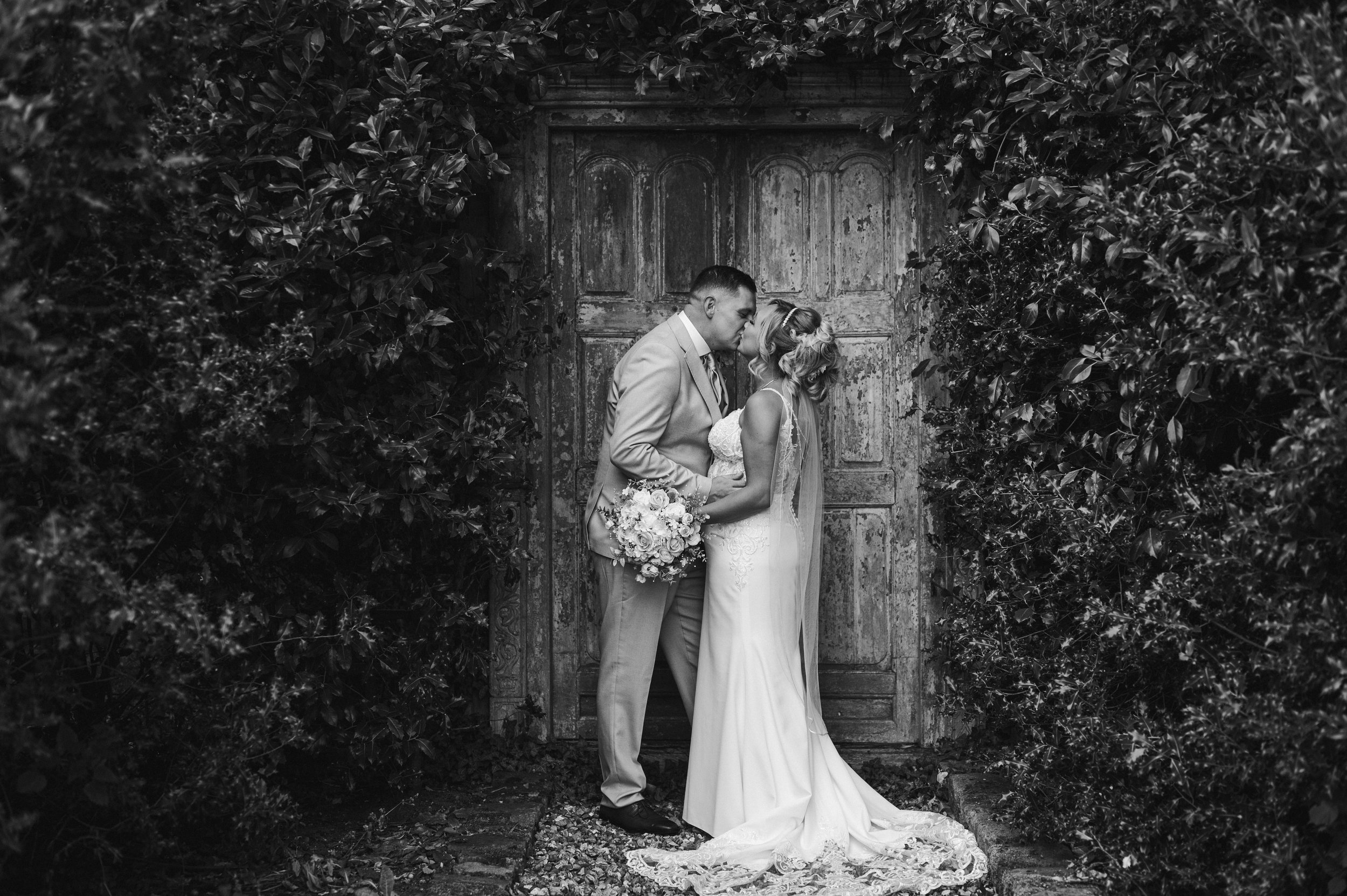 The Love Shack - black and white image of groom and bride in wedding attire, looking at each other and smiling in front of a wooden door. 