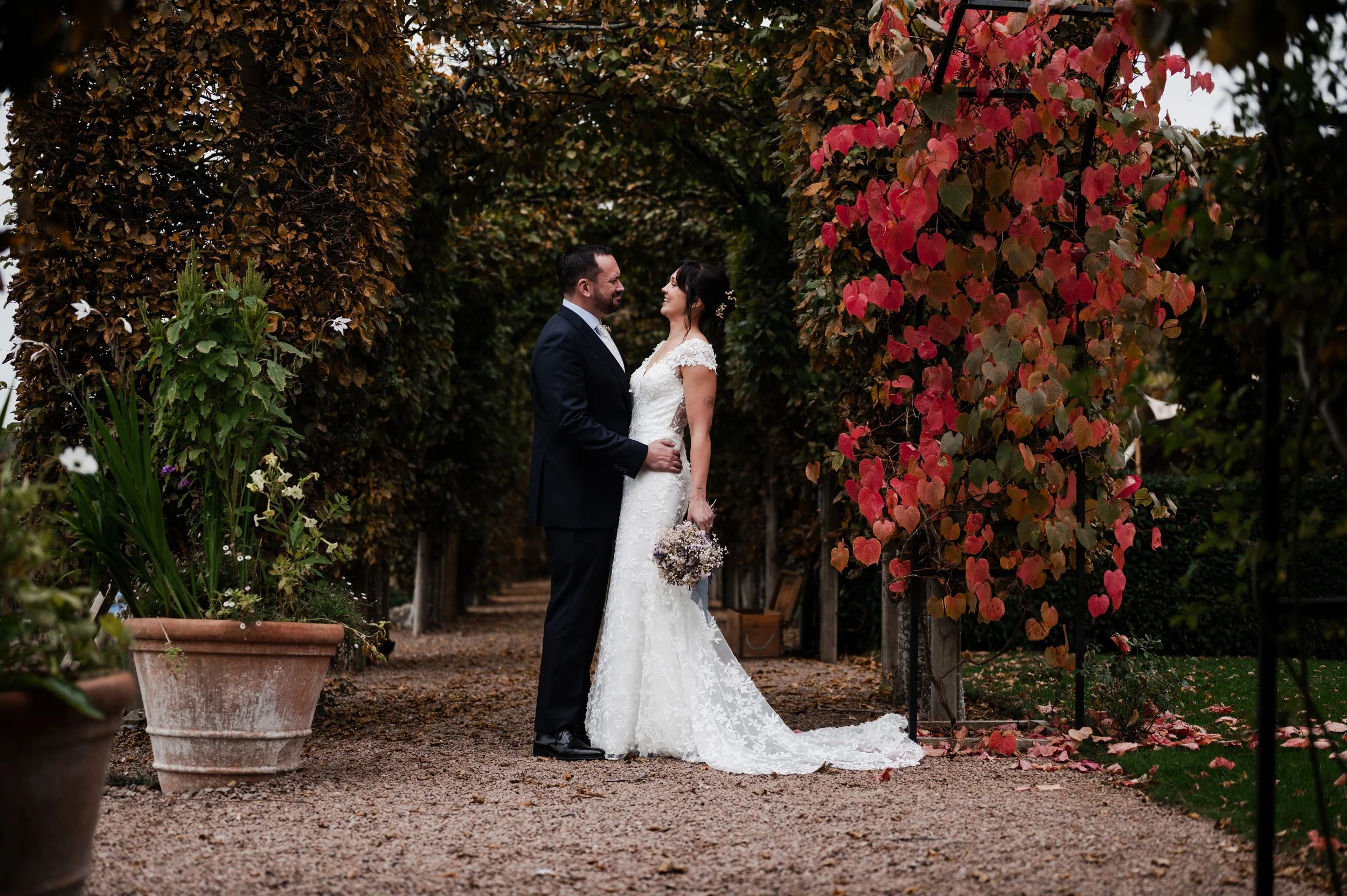 A newlywed couple standing close and gazing at each other in a garden path surrounded by autumn-colored foliage at pauntley court, Gareth roy photography