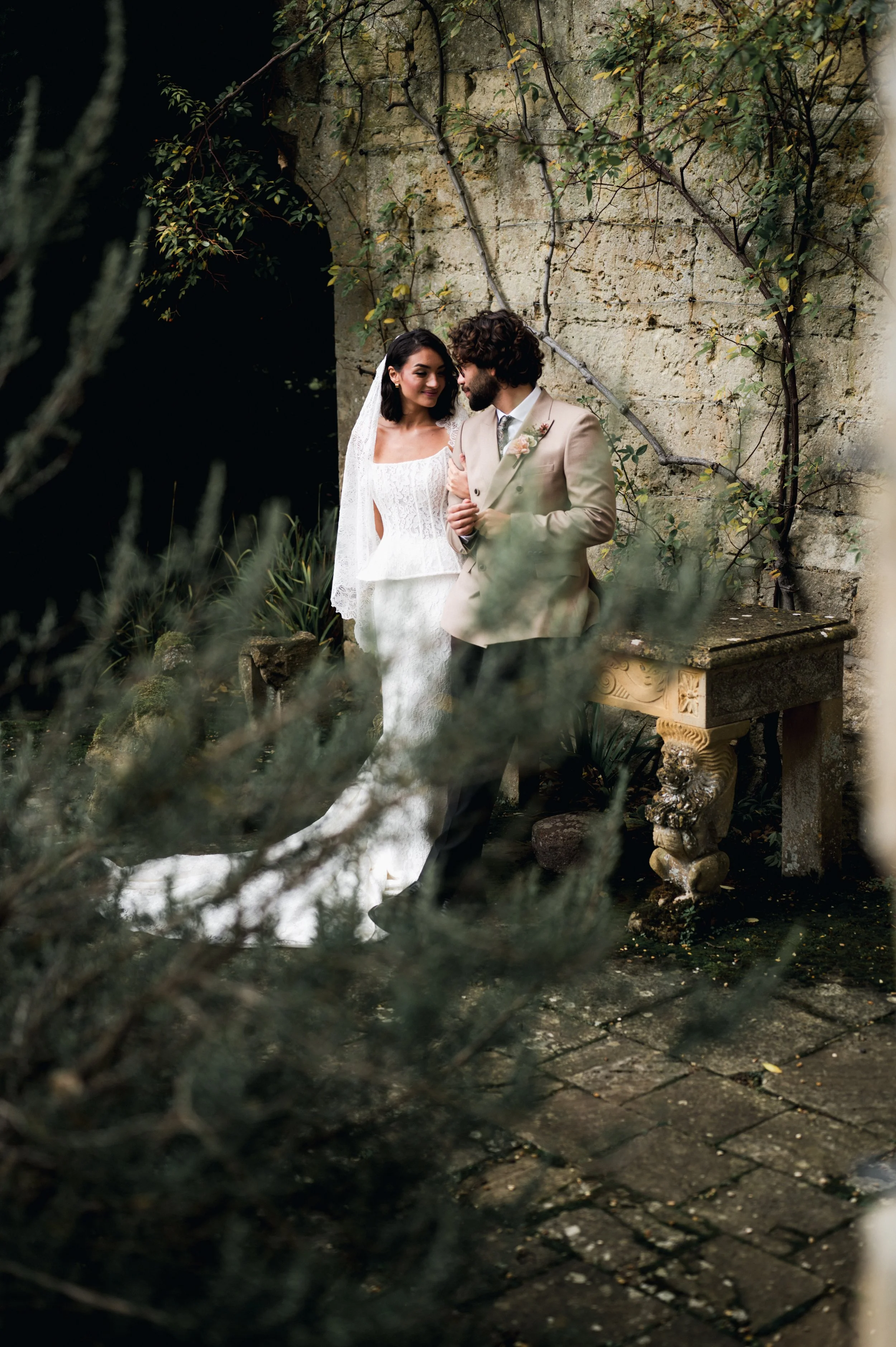 A bride and groom standing close together outdoors against a stone wall with climbing vines, surrounded by greenery, during a wedding photoshoot at sudeley castle, Gareth roy photography