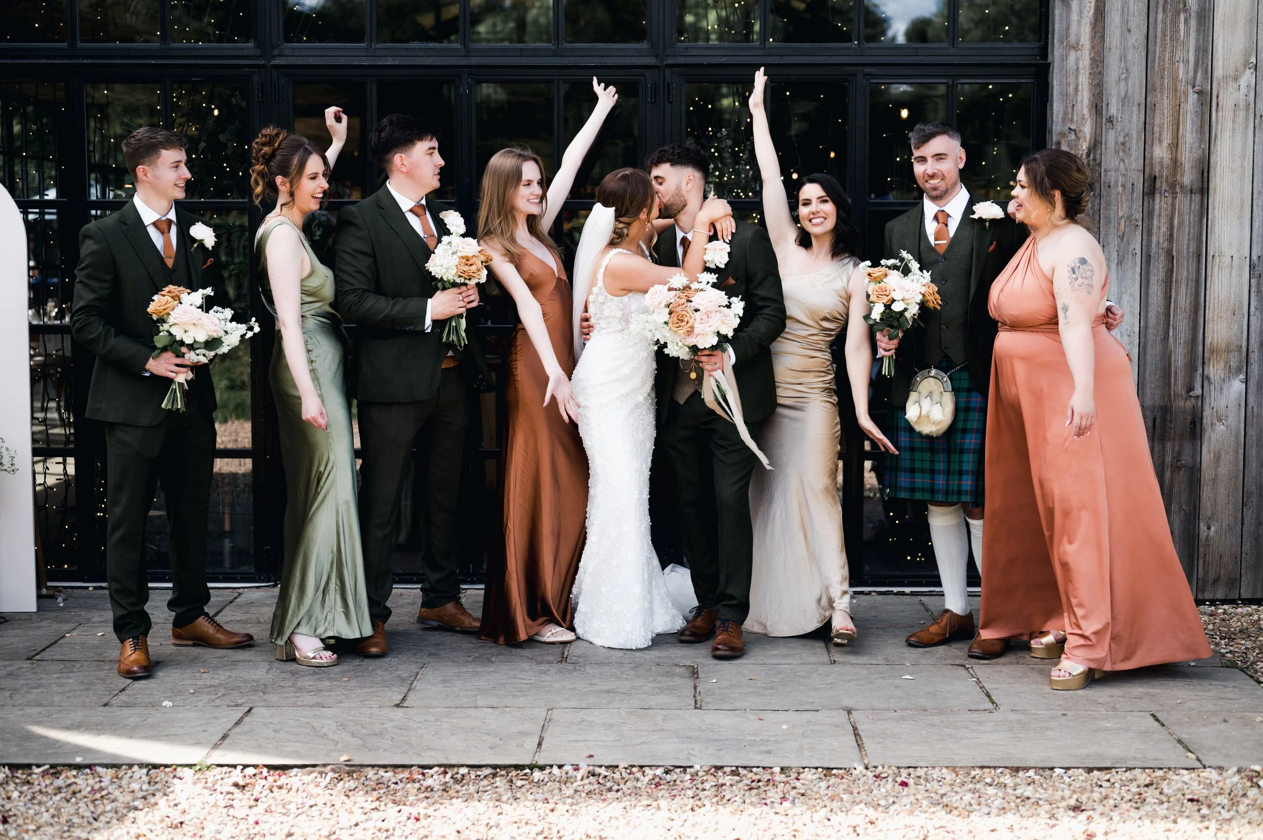 Group of wedding guests and bride and groom celebrating in front of a building with glass doors, holding bouquets and raising their hands hidden river cabins, Gareth roy photography
