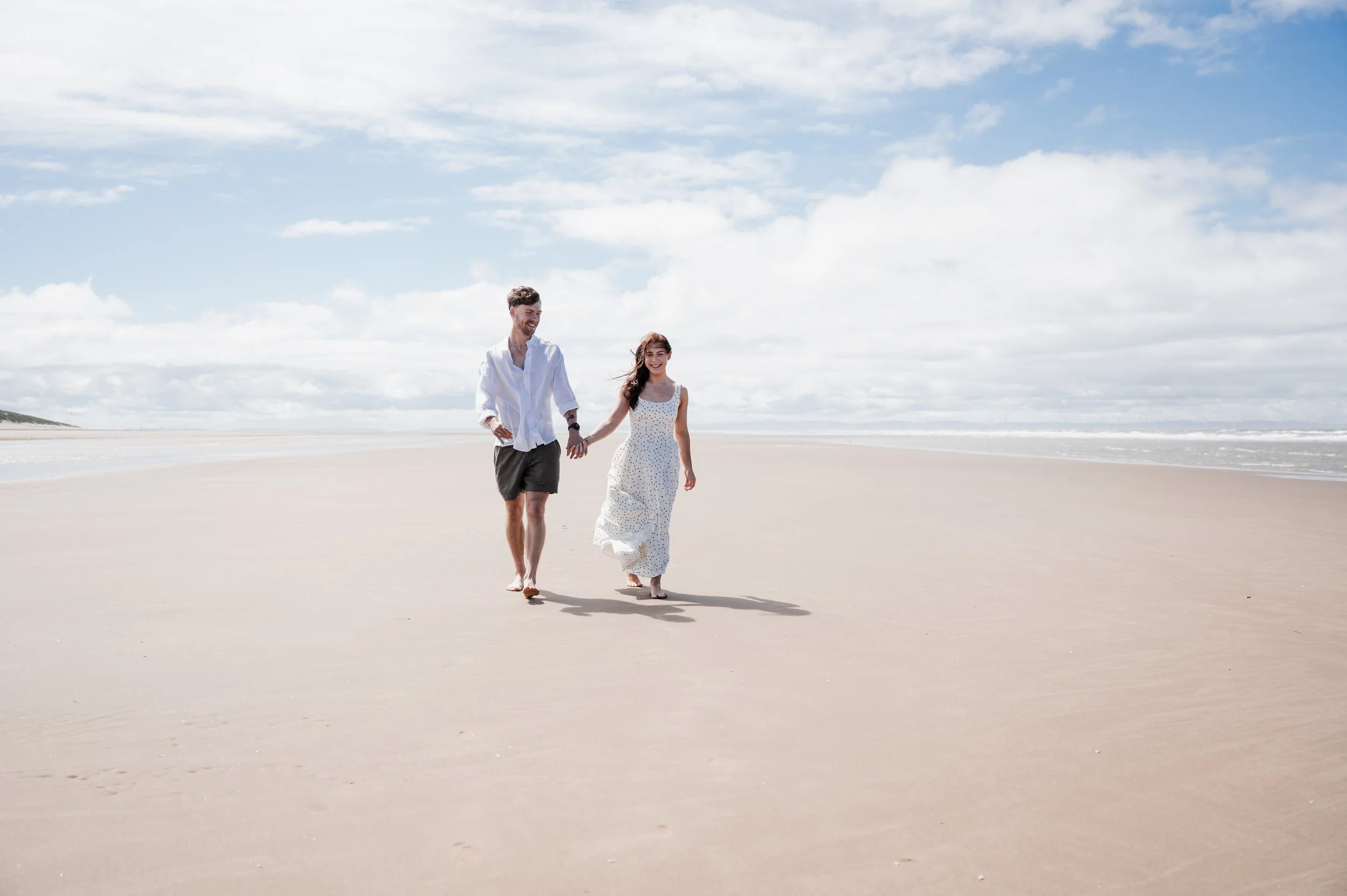 Formby Beach Engagement - Man and woman wearing white and black clothes on a beach , holding each other's hands and smiling