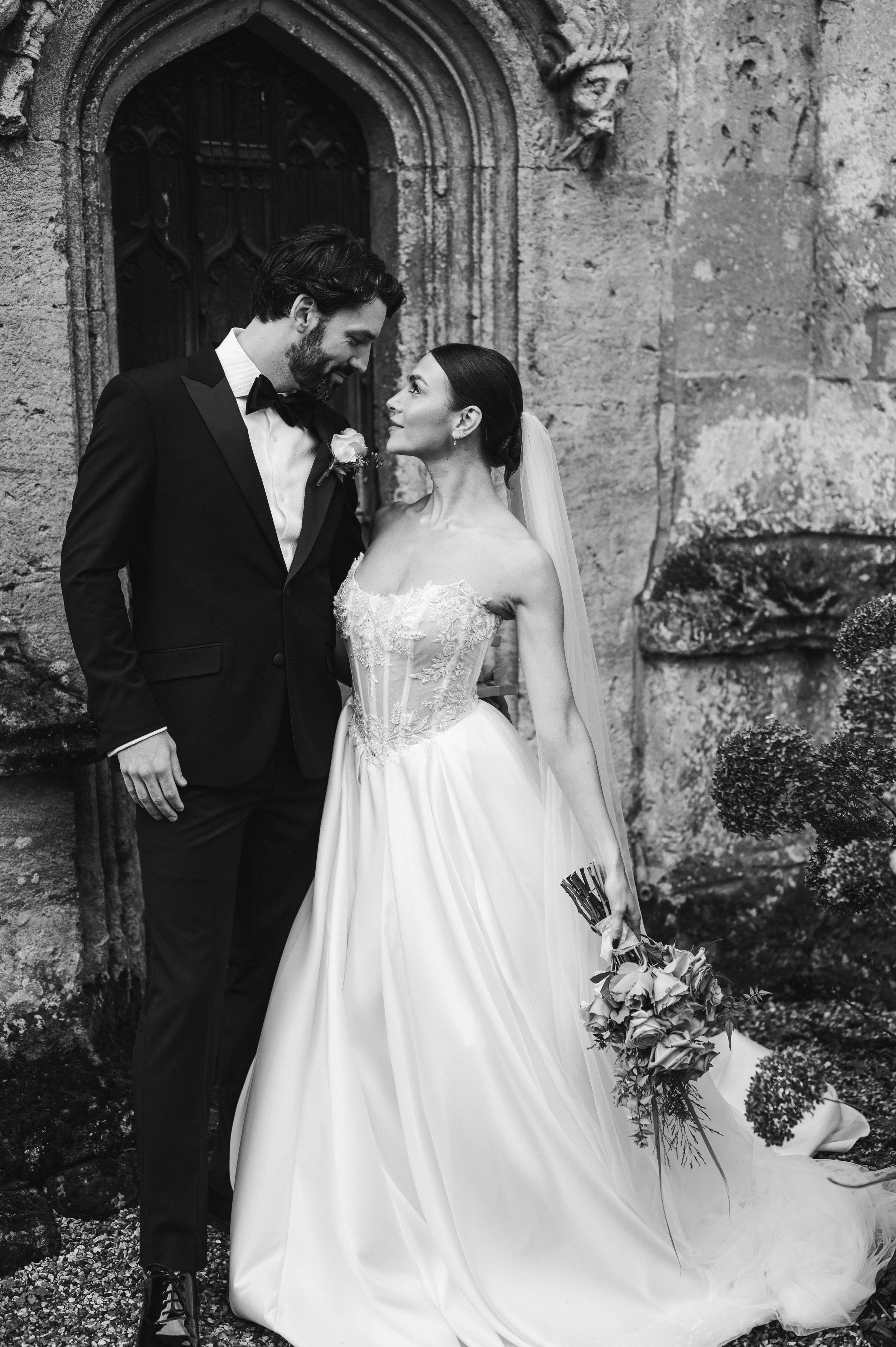 Black and white image of bride and groom in wedding attire looking at each other outside a stone doorway