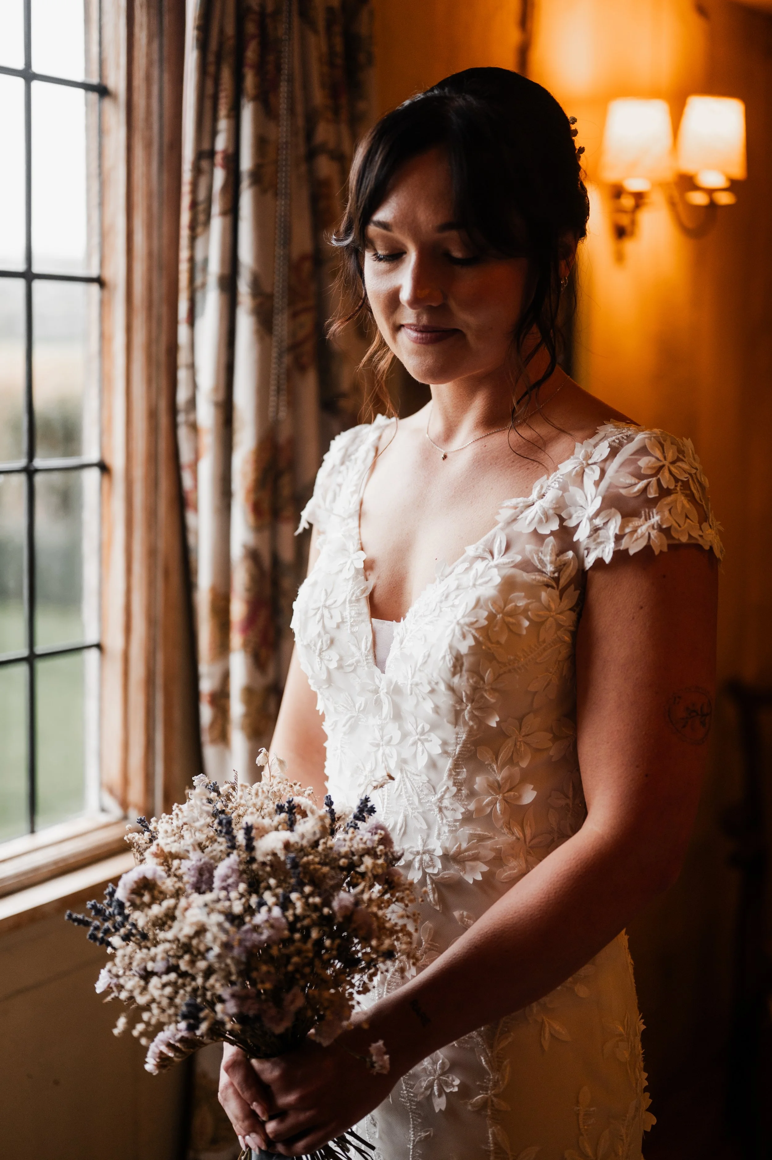 A woman on her wedding day in a white lace dress with floral details, holding a bouquet of dried flowers, standing inside near a window and looking down softly pauntley court, Gareth roy photography