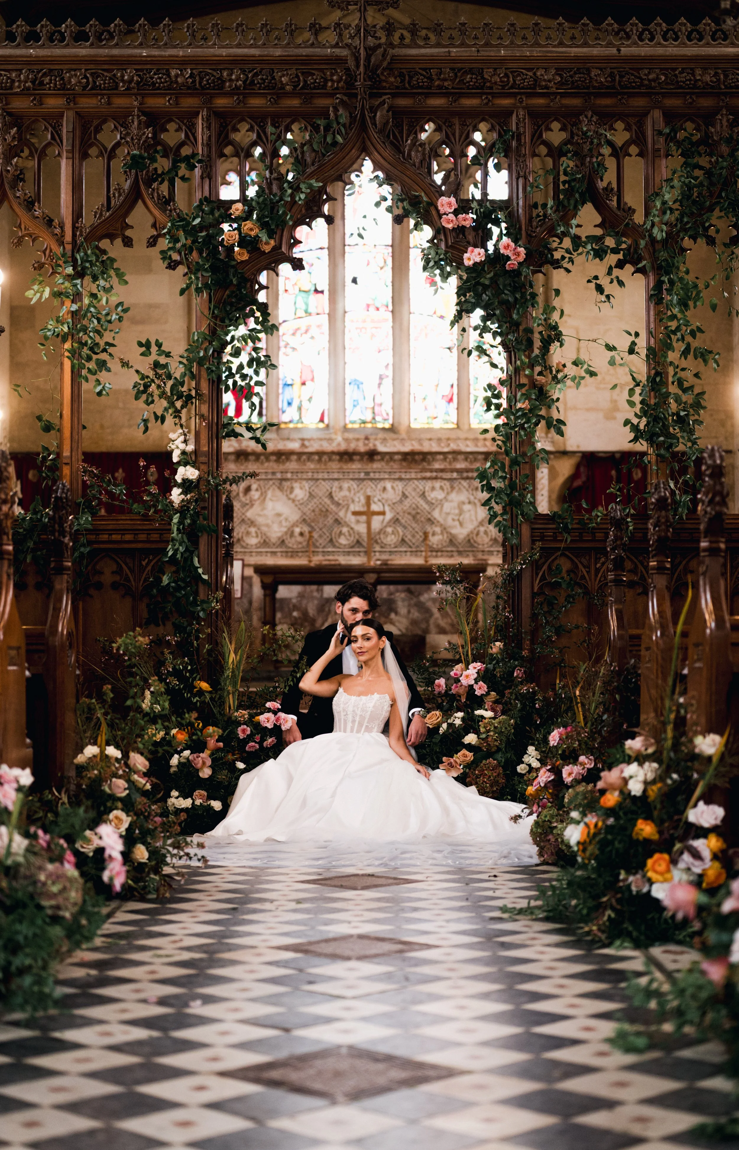 Bride and groom in wedding attire, sat on wedding aisle steps, surrounded by flowers and sat under an archway