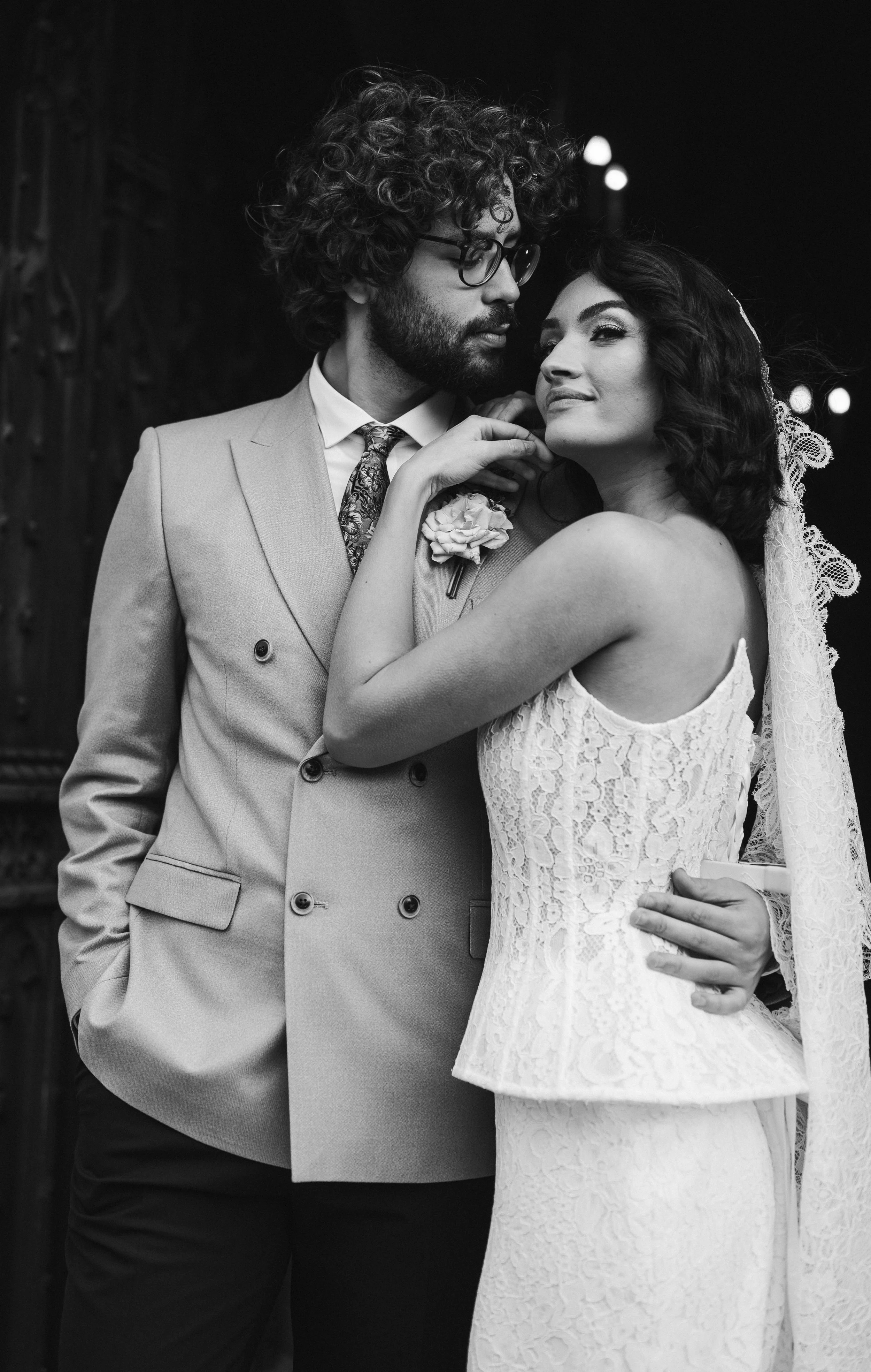 A black and white photo of a bride and groom, with the groom wearing glasses and a double-breasted suit, and the bride wearing a lace wedding dress and veil, standing close together at night with blurred lights in the background at sudeley castle