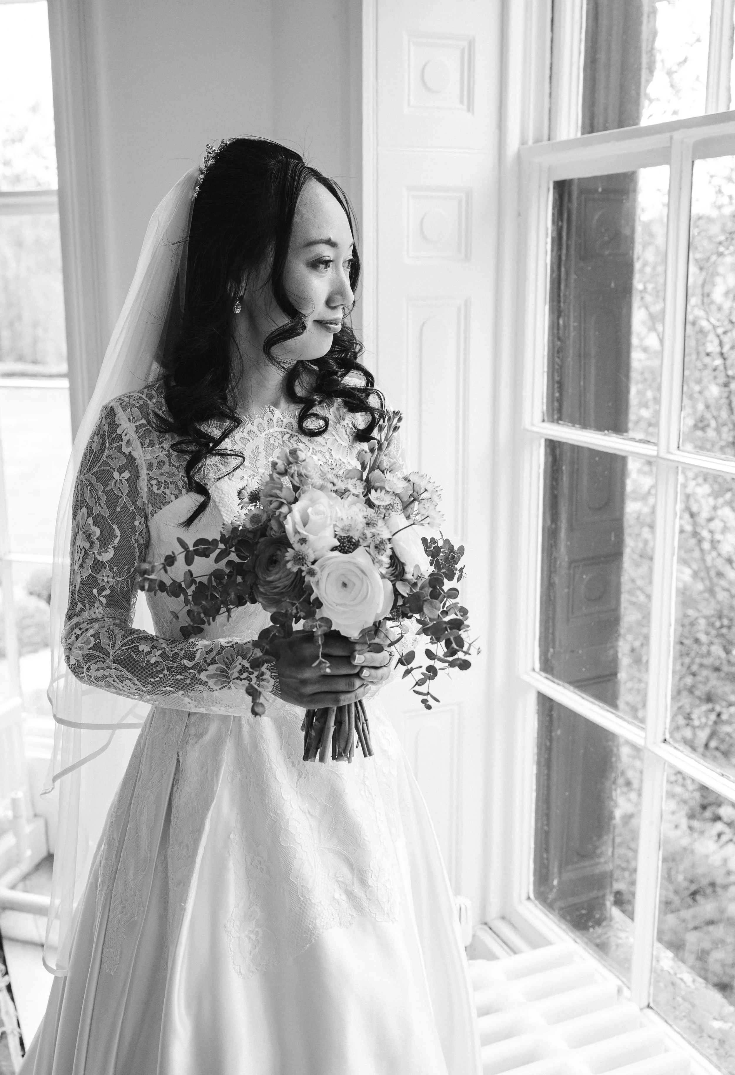 Black and white image of a bride in a white lace dress, looking out the window and holding a bouquet
