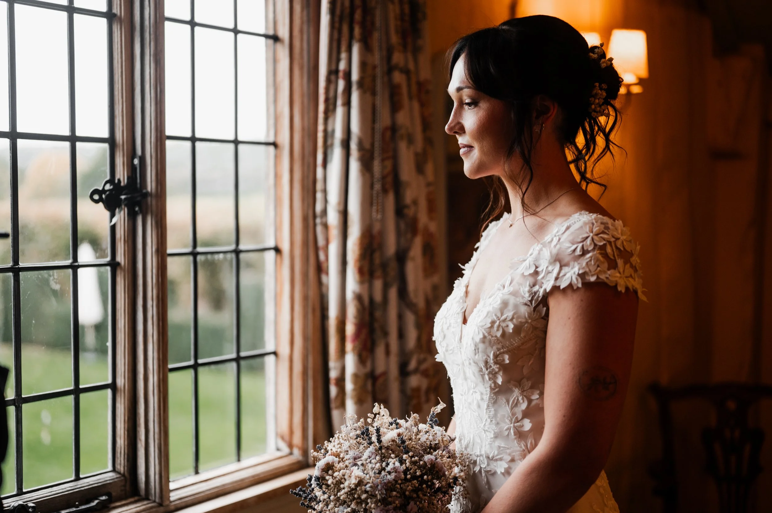 A bride standing by a window in a cozy room, holding a bouquet of flowers, wearing a wedding dress with floral details, looking outside thoughtfully pauntley court, Gareth roy photography