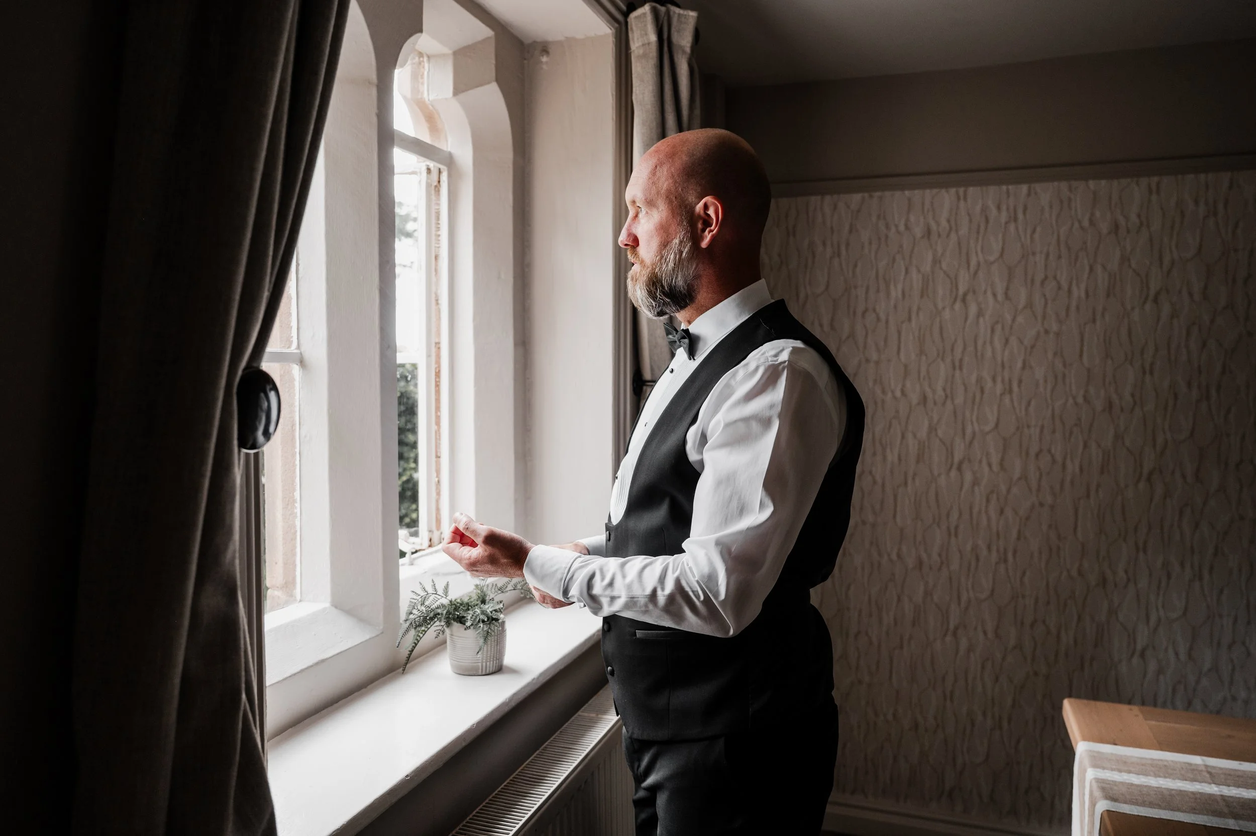Peckforton Castle - groom getting ready in wedding attire while looking outside a bright window