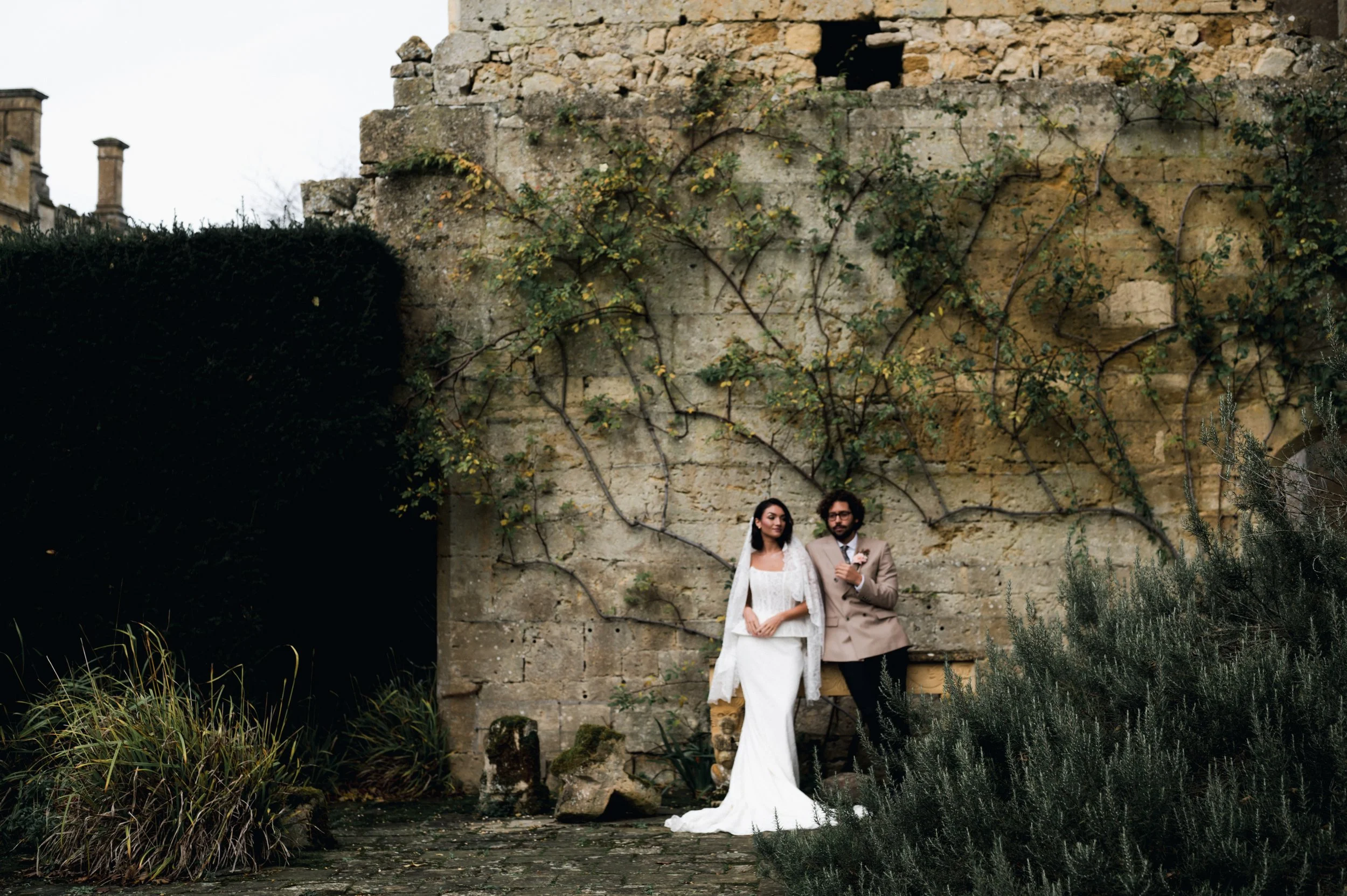 A bride and groom standing outdoors against an old stone wall with vines, surrounded by greenery, on their wedding day at sudeley castle, Gareth roy photography