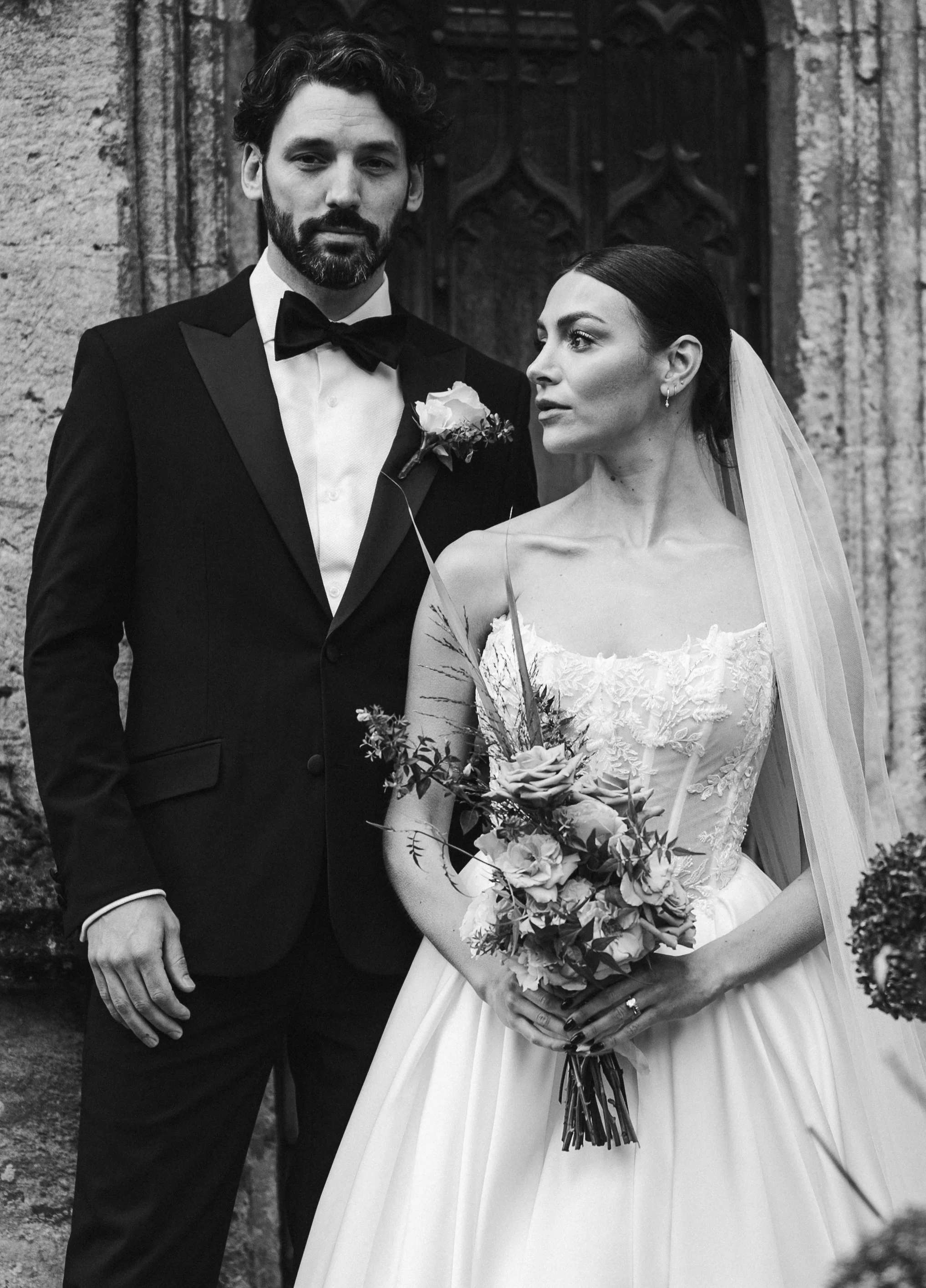 A black and white photo of a bride and groom standing outdoors, with the groom in a tuxedo and the bride in a wedding dress holding a bouquet at sudeley castle, Gareth roy photography
