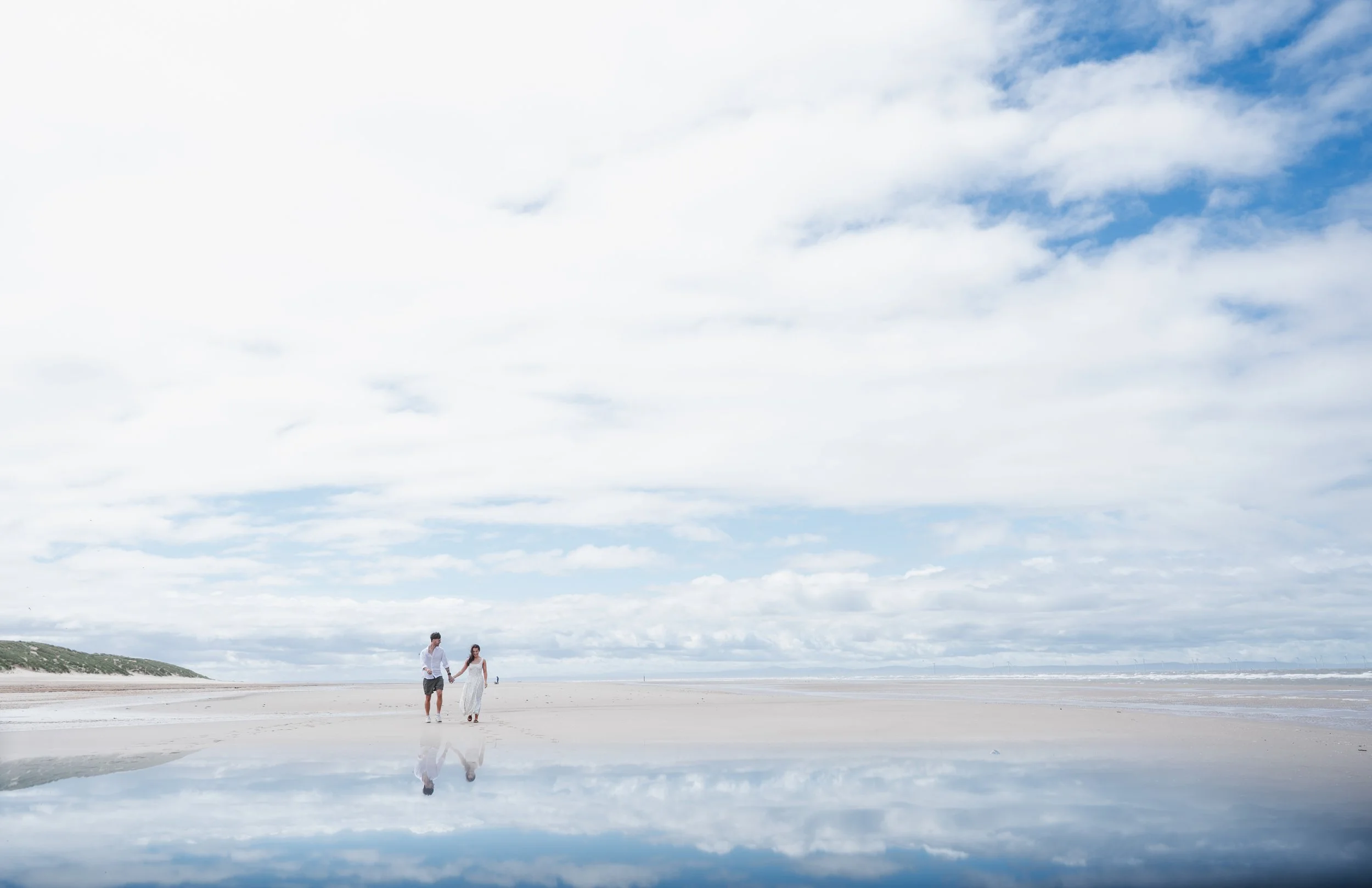 Formby Beach Engagement - Man and woman wearing white clothes on a beach, photographed from a distance with cloud on a blue sky above