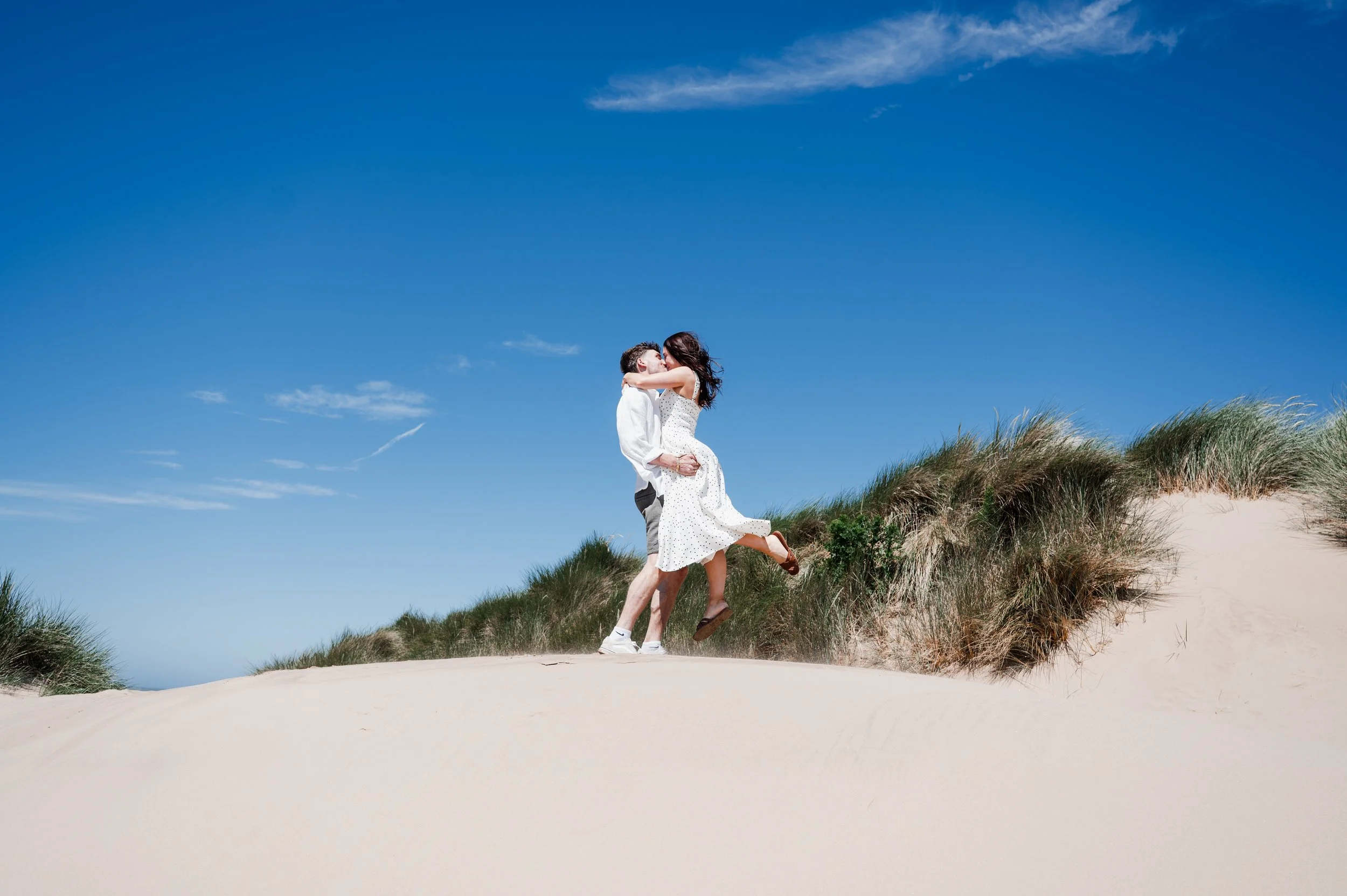 Formby Beach Engagement - Man and woman wearing white clothes stood on sand dunes while woman is lifted and spun around. 