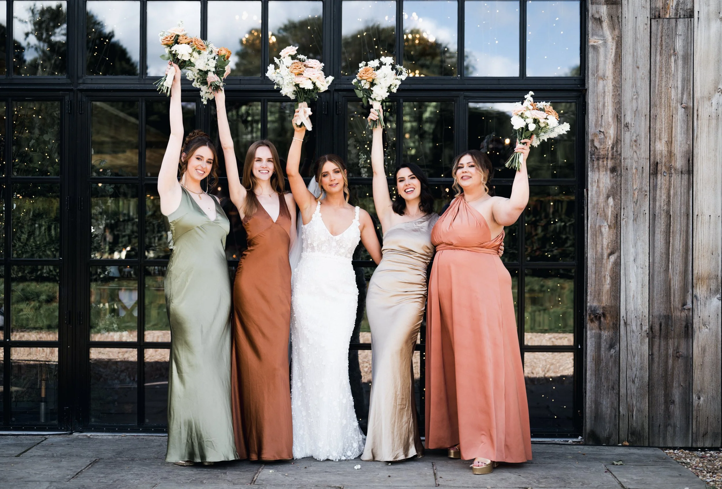 Five women in elegant gowns, standing in front of a glass window with string lights, celebrating at a wedding. They are holding bouquets raised in the air, smiling happily. hidden river cabins, Gareth roy photography