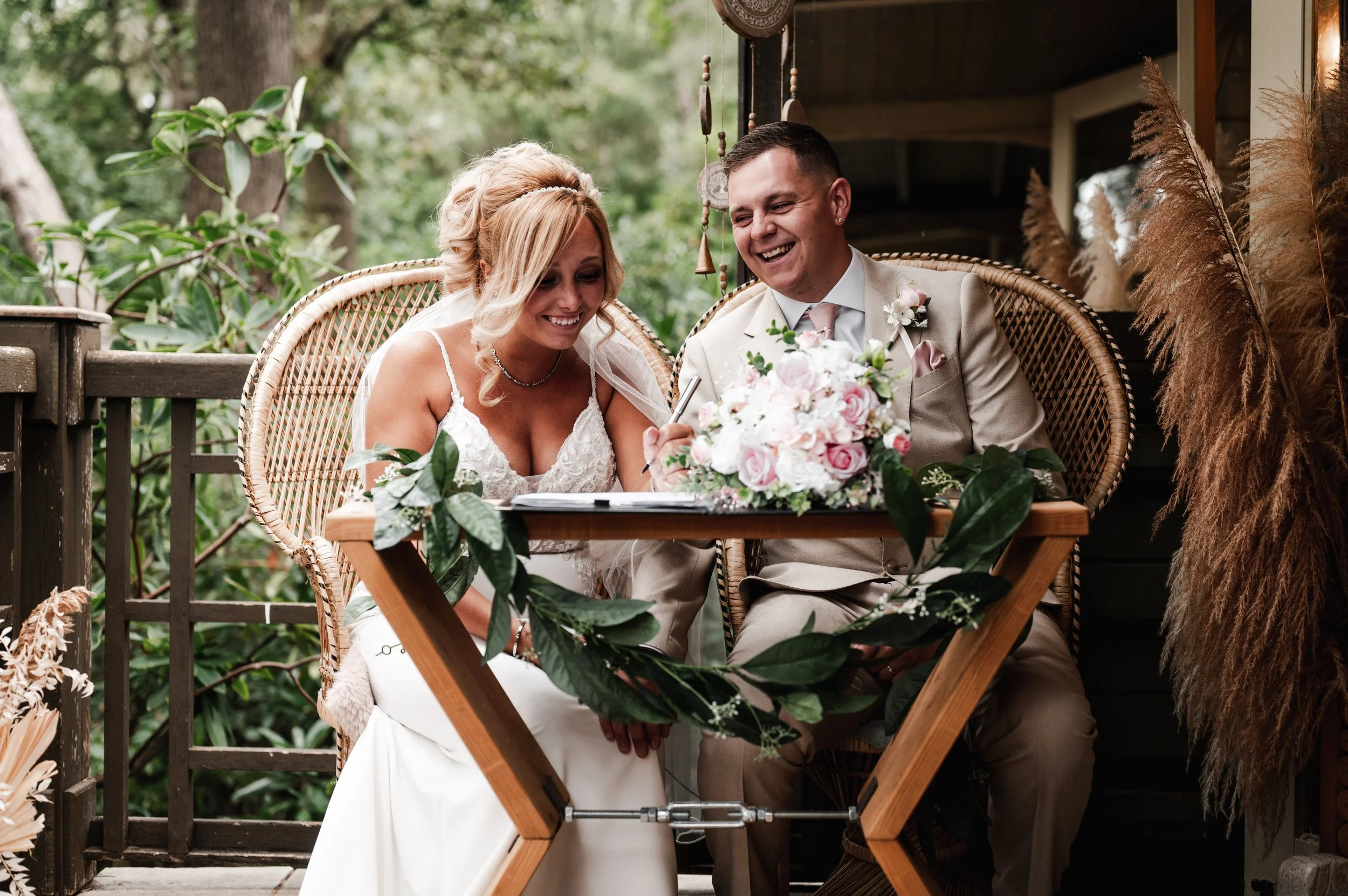 The Love Shack - Groom and bride in wedding attire, sat on wooden chair signing a register and laughing. 