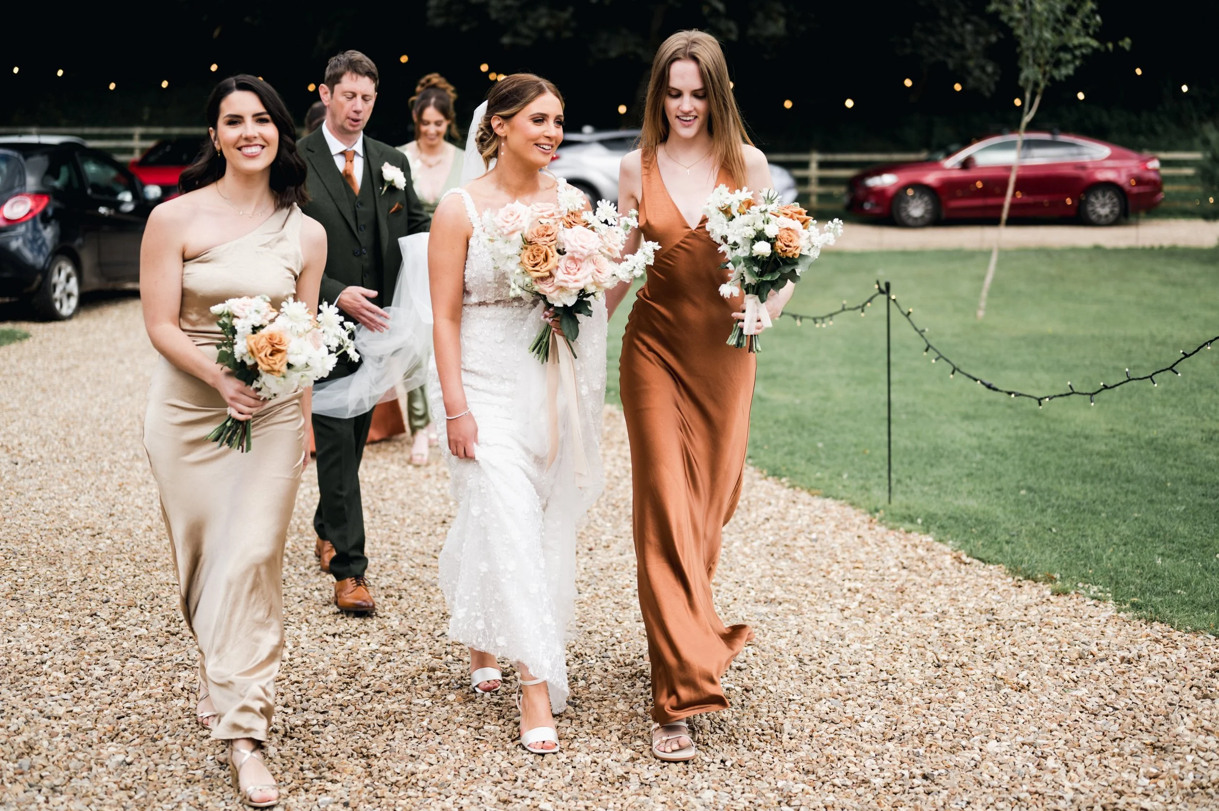 Group of five people, two women in wedding dresses, walking outdoors on a gravel path, holding bouquets of flowers hidden river cabins, Gareth roy photography