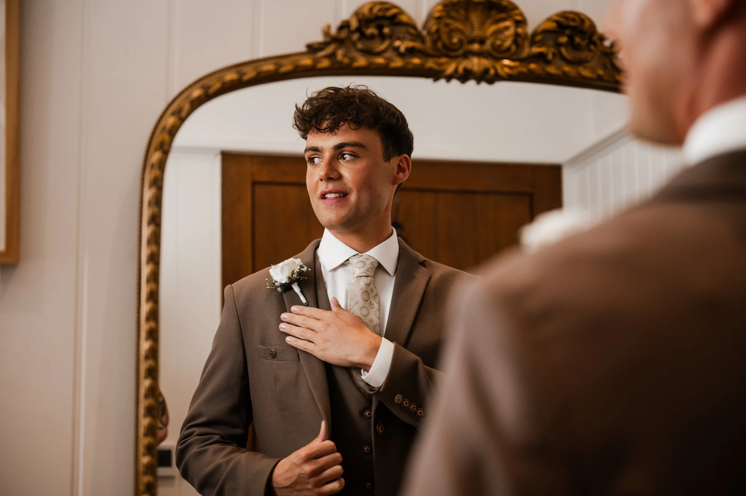 Groom in brown suit getting ready in a mirror and looking off to the side