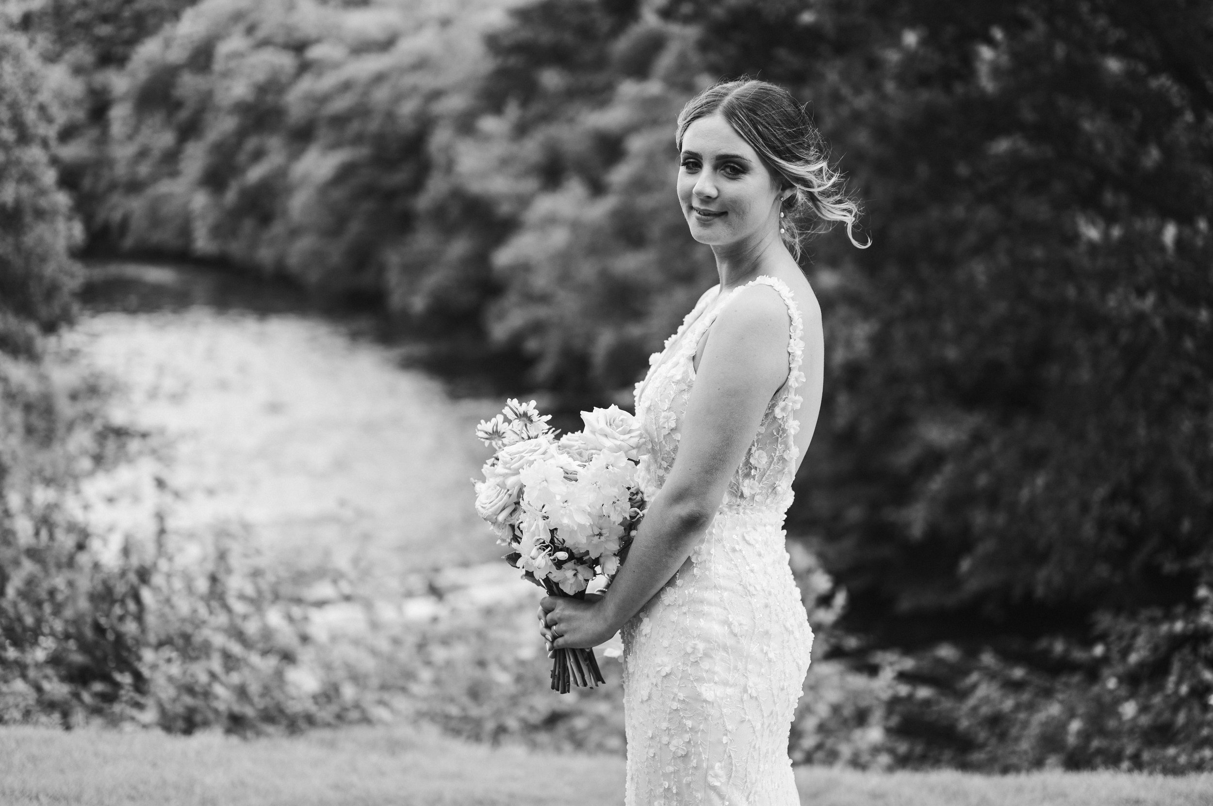 A woman in a wedding dress holding a bouquet of flowers outdoors with trees and a body of water in the background hidden river cabins, Gareth roy photography