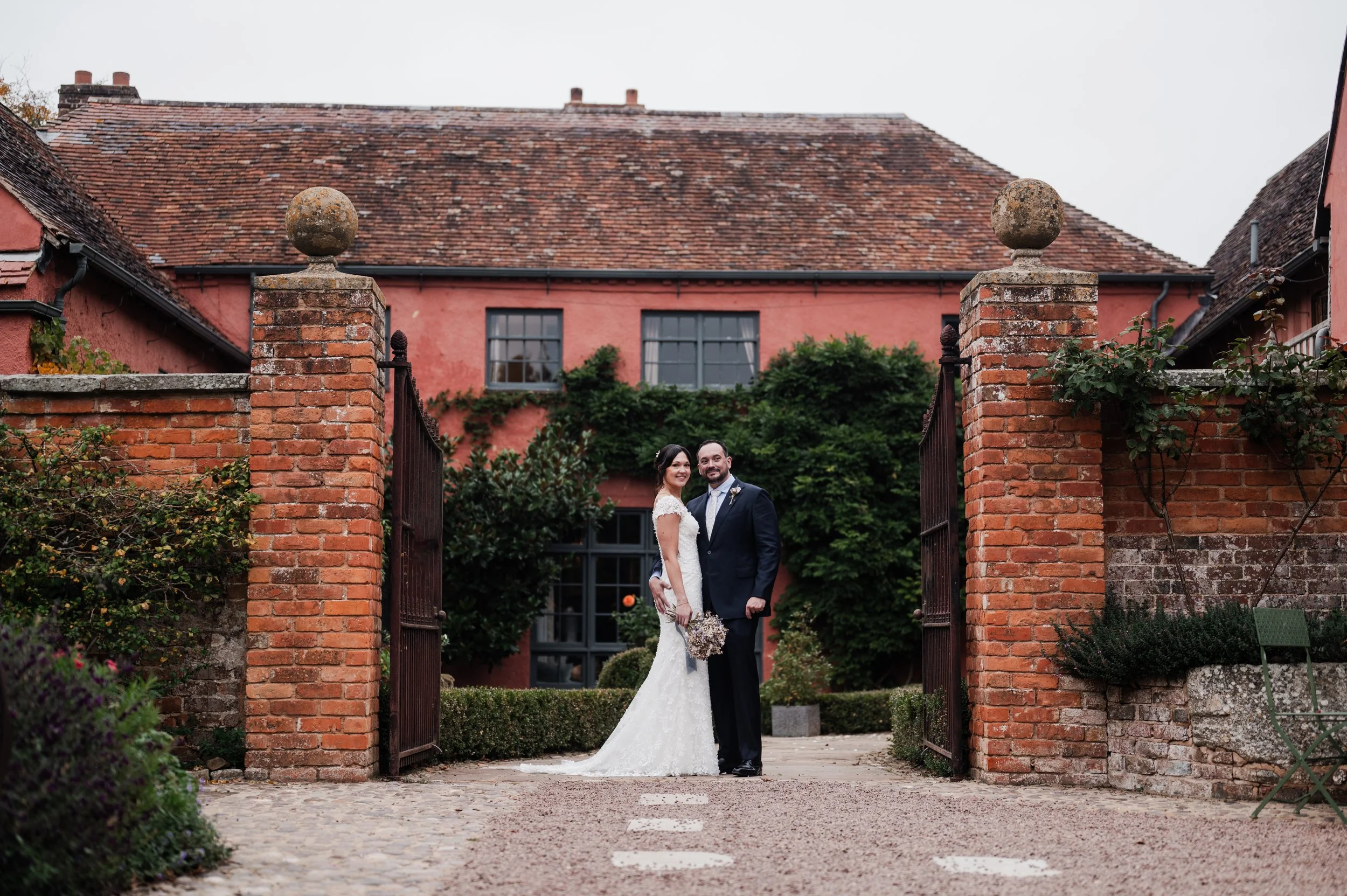 A bride and groom standing together behind an open brick gate in a garden, with a red building and greenery in the background pauntley court, Gareth roy photography