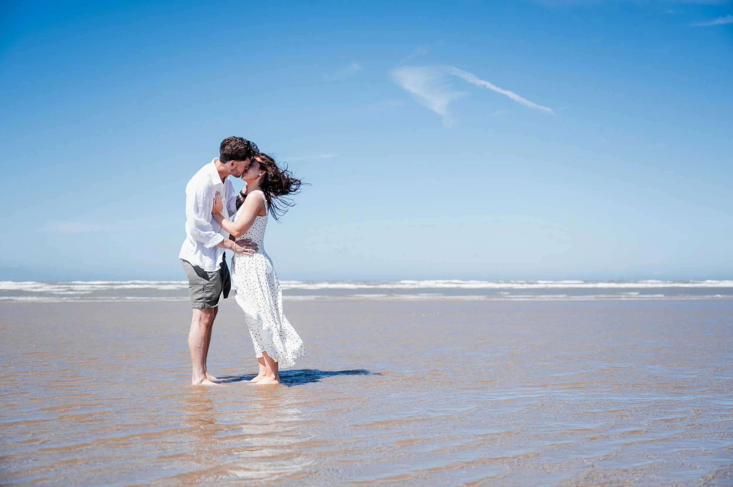Formby Beach Engagement - Man and woman wearing white clothes stood in shallow water on a beach while kissing