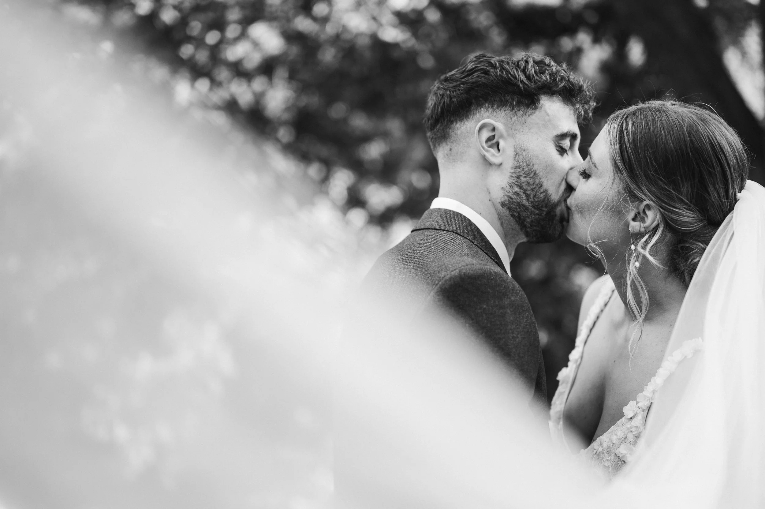 A black-and-white photograph of a newlywed couple kissing outdoors, blurred foreground, trees in the background hidden river cabins, Gareth roy photography