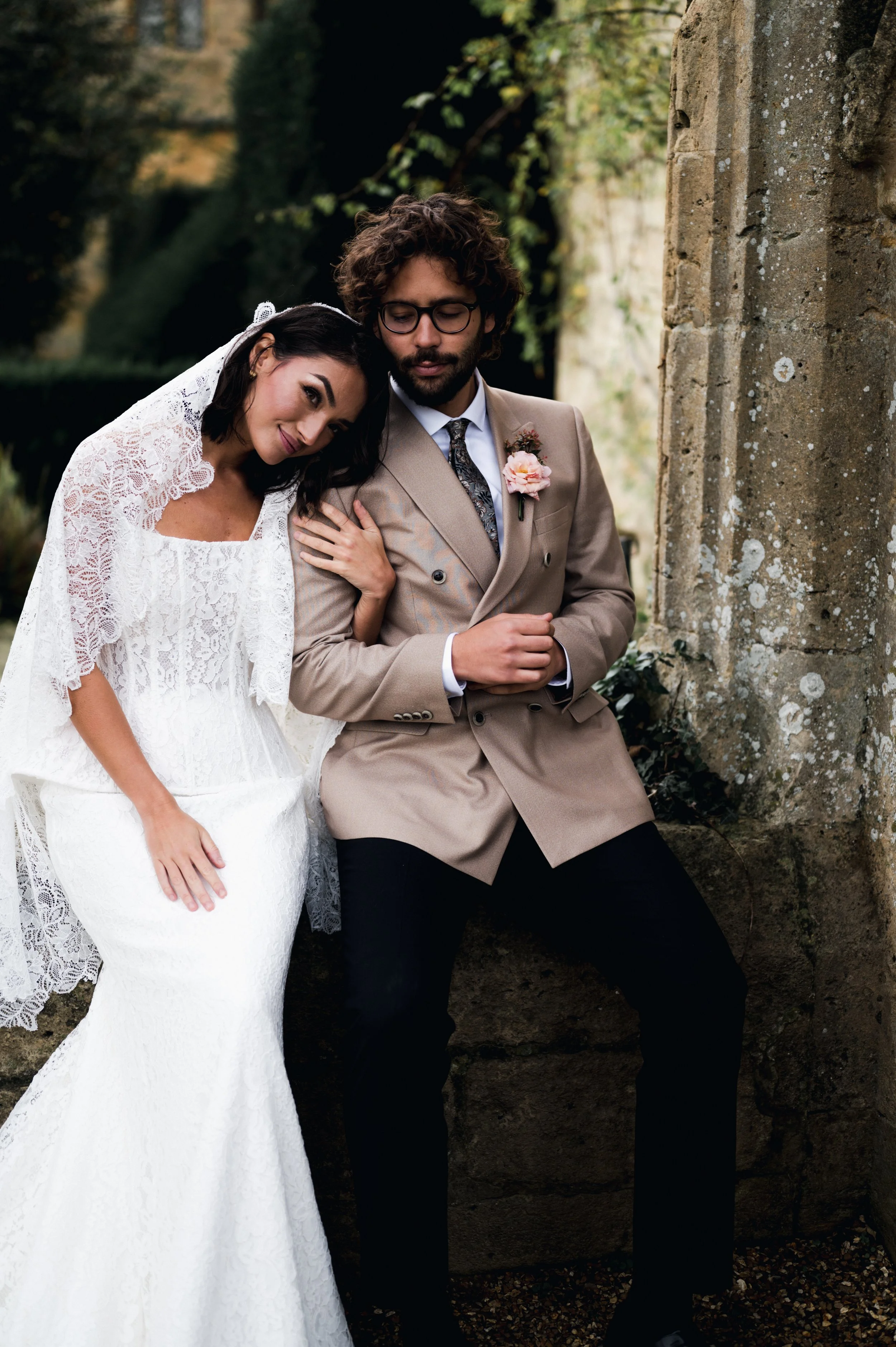 A bride and groom sitting outdoors near stone architecture, with the bride resting her head on the groom's shoulder, both dressed in wedding attire at sudeley castle, Gareth roy photography