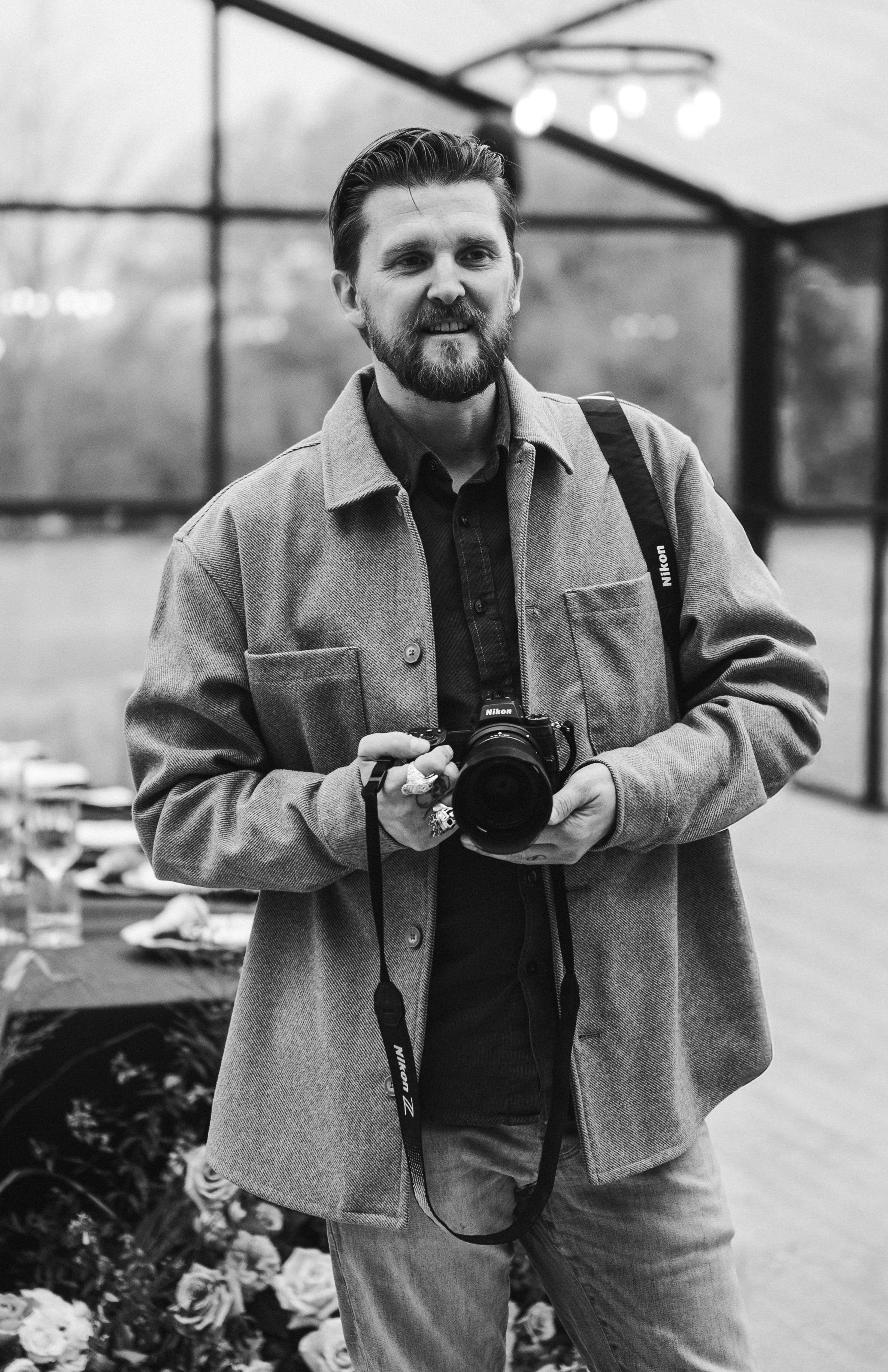 A man with a beard and mustache holding a camera, standing in front of a flower display in a greenhouse or flower shop, smiling at the camera.