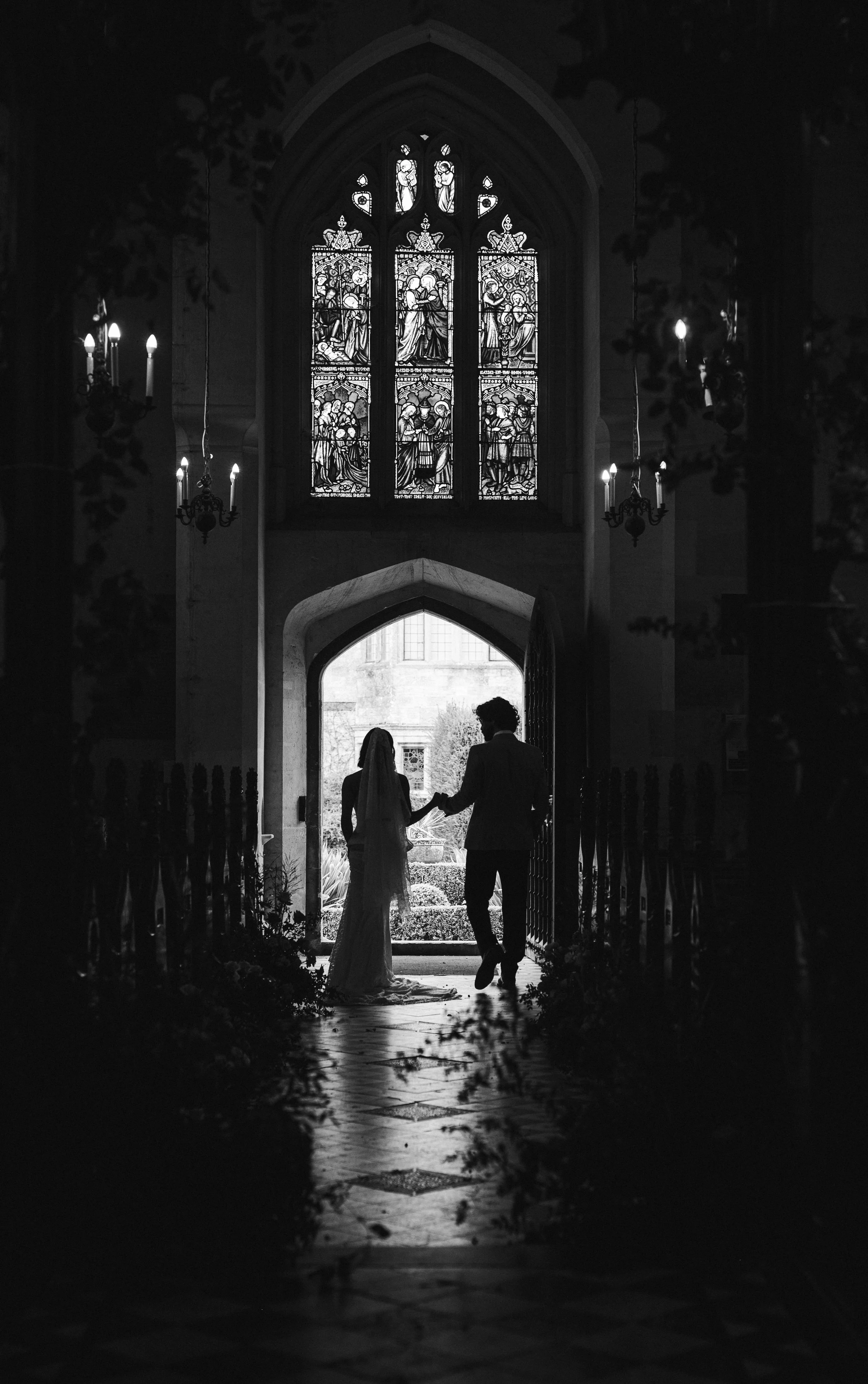 Silhouettes of a bride and groom holding hands in a church aisle, with stained glass windows above them, black and white photo at suedley castle