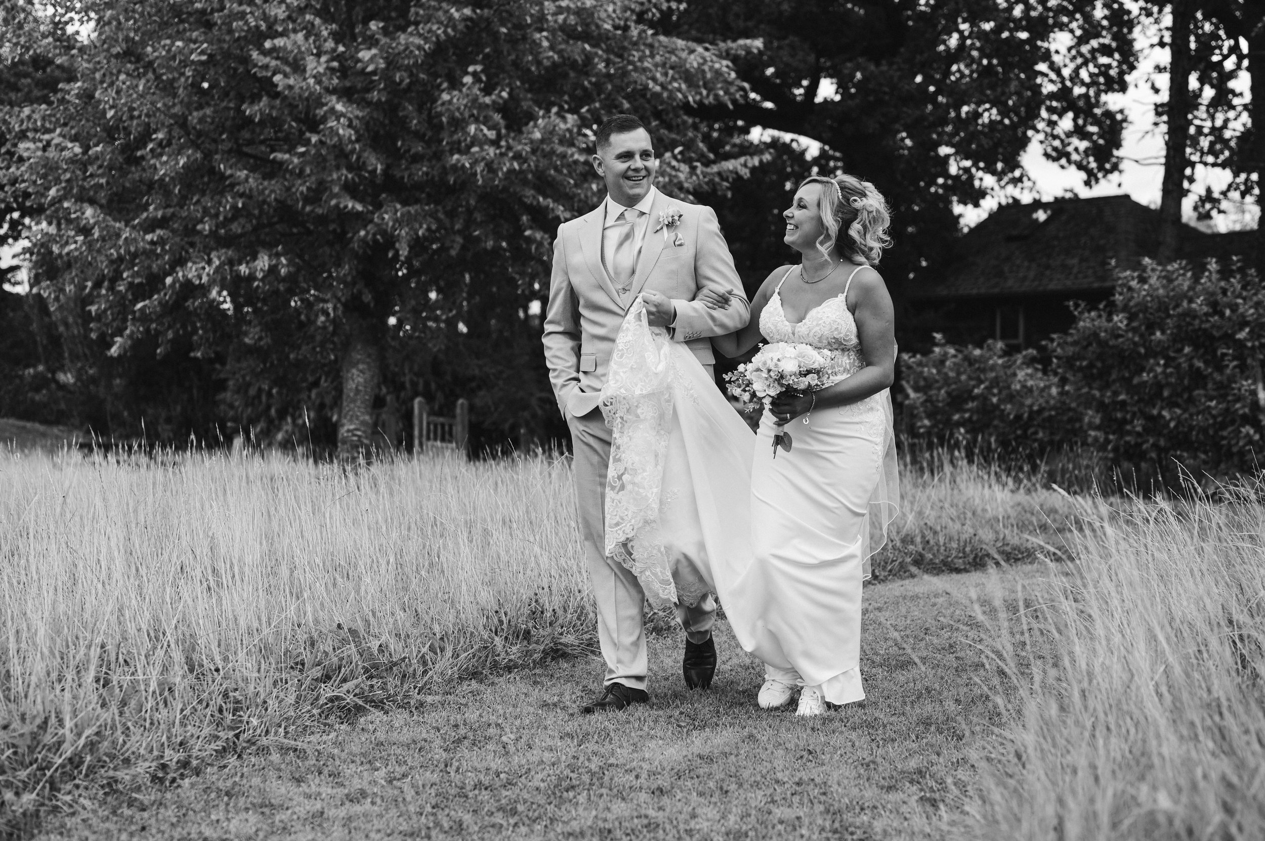 The Love Shack - black and white image of groom and bride in wedding attire, looking at each other and smiling while walking down a grassy path. 