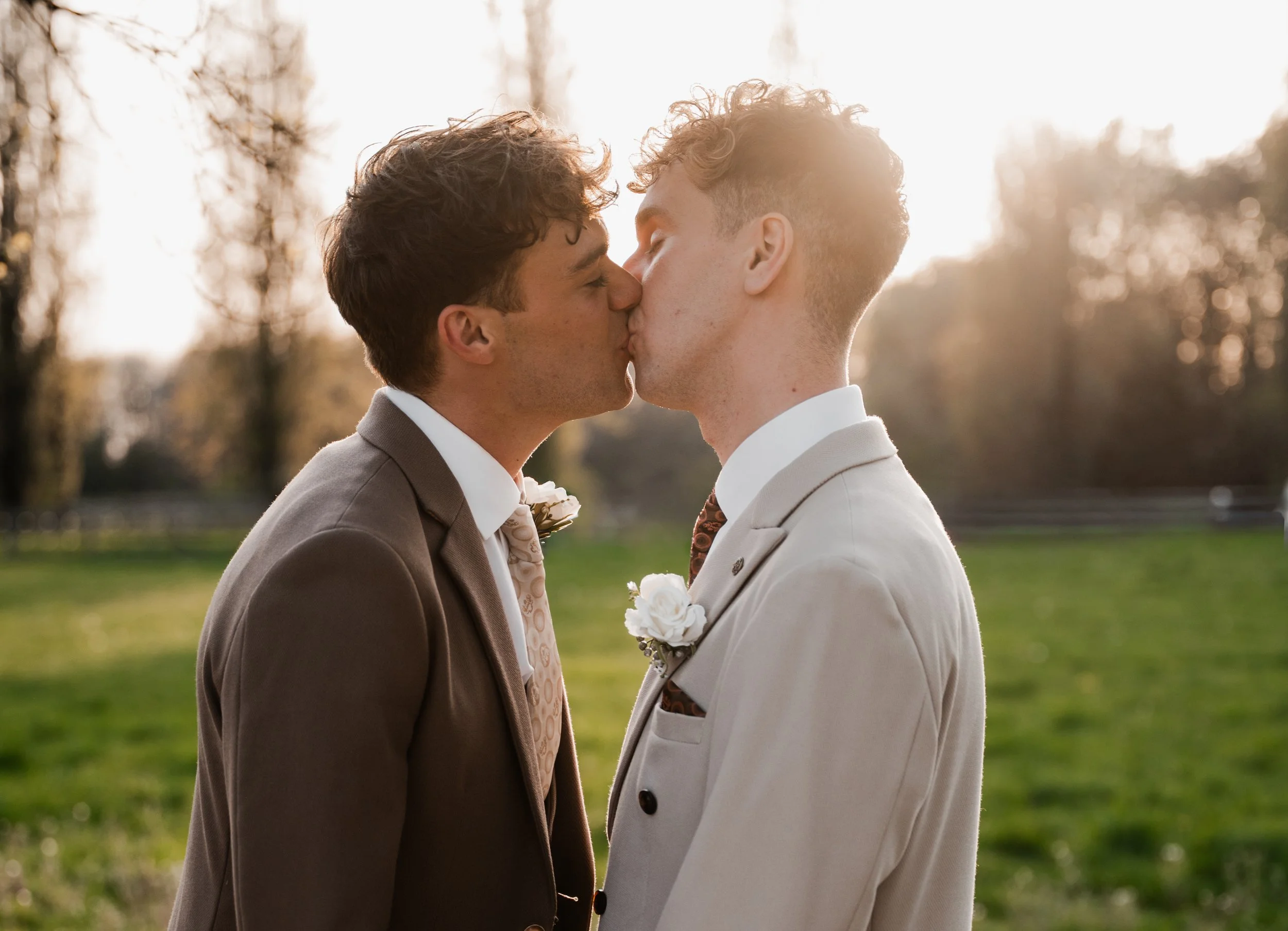 Two grooms kiss in front of a sunset and in venue gardens