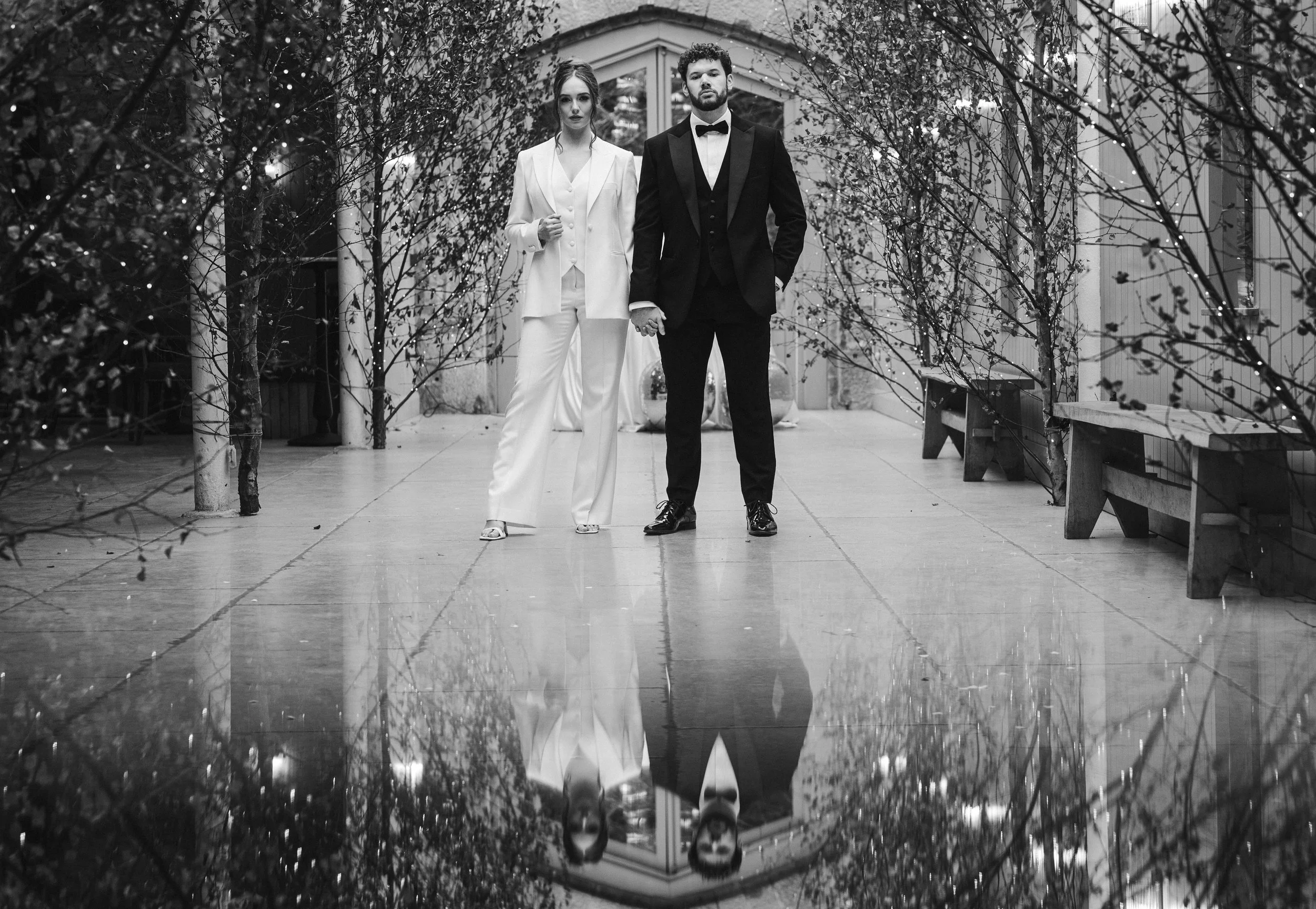Wedding Photographer Cheshire - Black and white image of bride and groom stood in a tree lined aisle with their reflections on the stone floor
