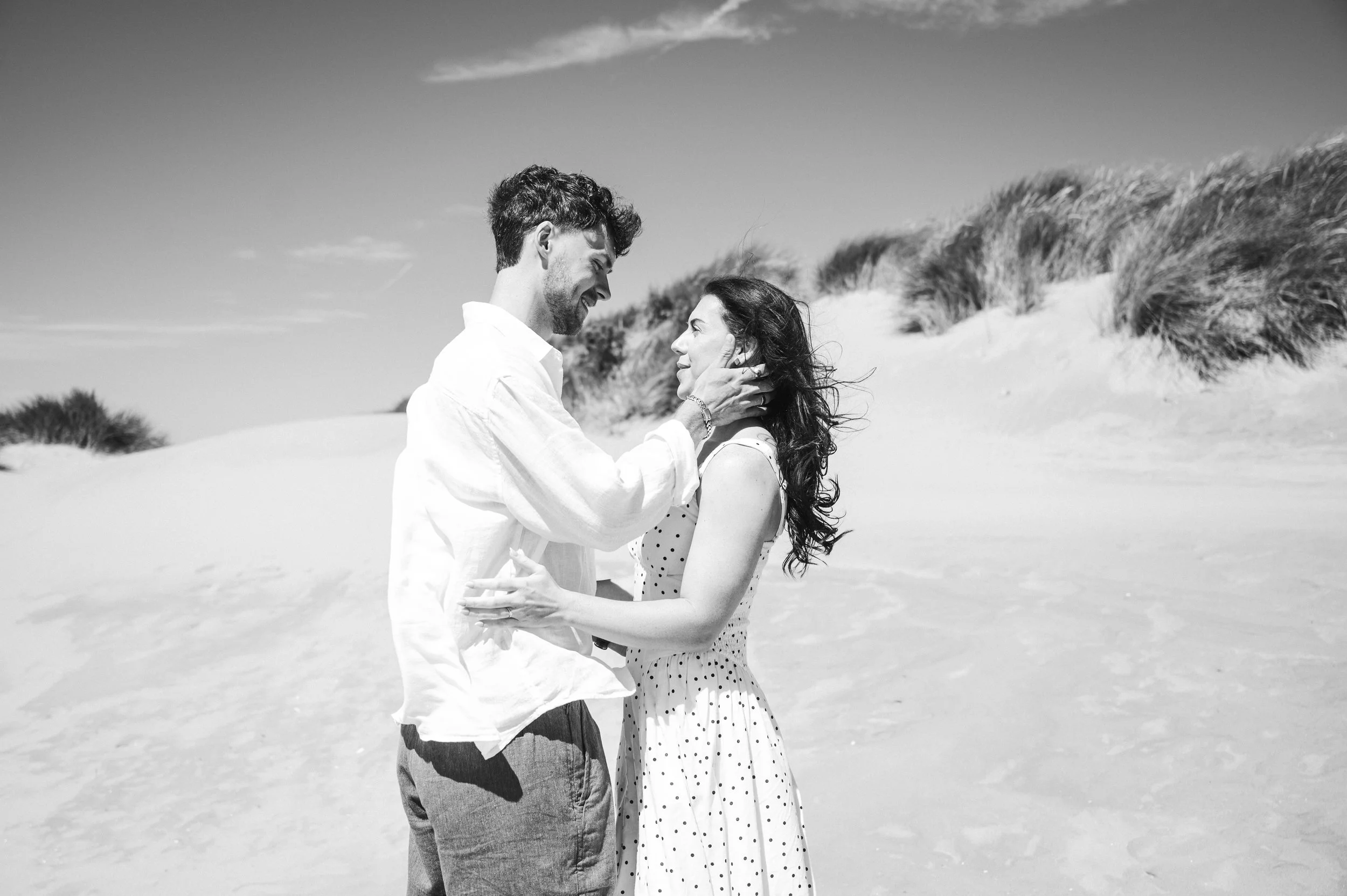 Formby Beach Engagement - black and white image of a man and woman wearing white clothes on a beach , holding each other and smiling in an embrace