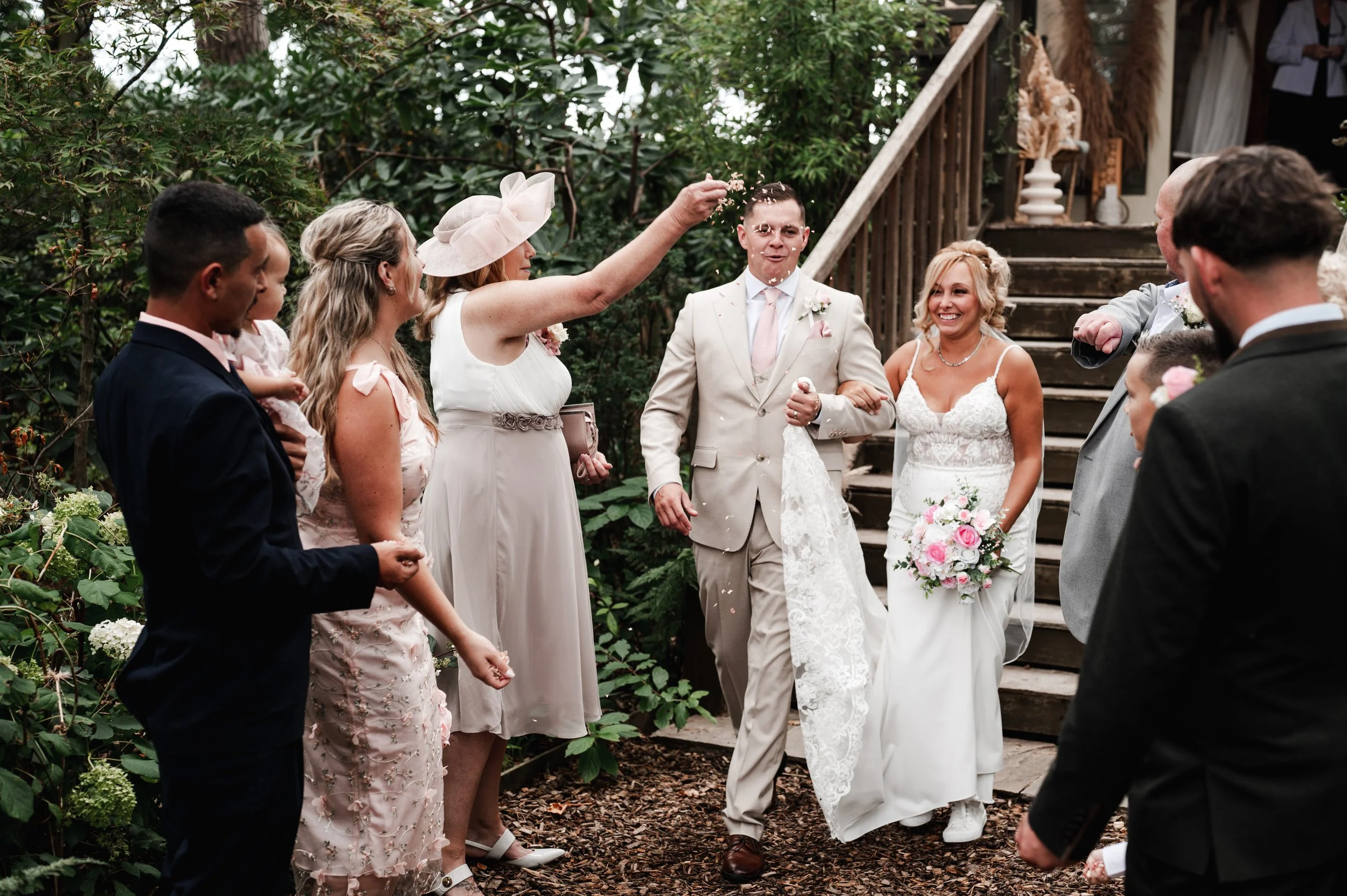 The Love Shack - Groom and bride in wedding attire, stood at bottom of a wooden staircase while guests throw confetti.
