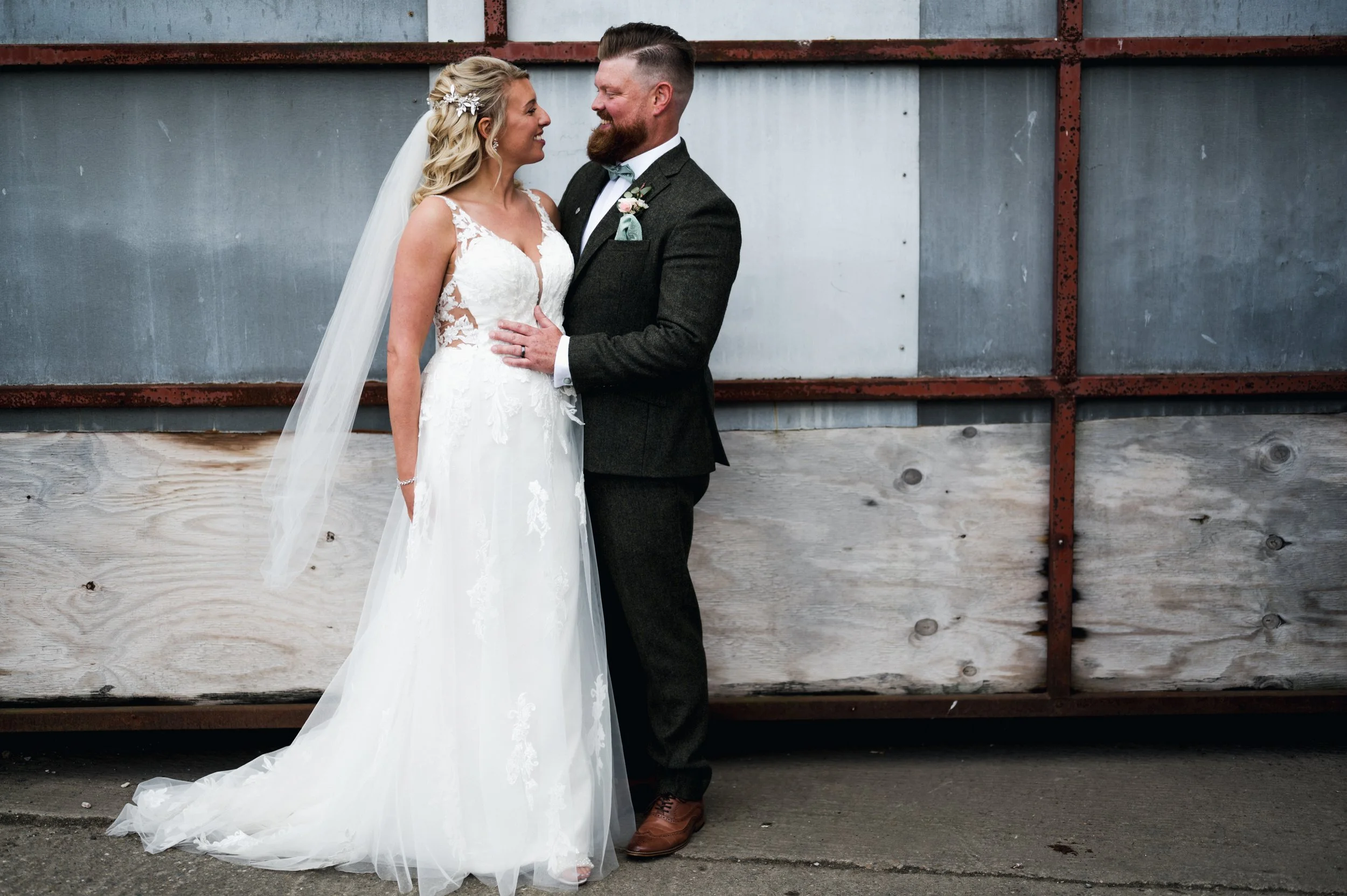 A bride and groom stand close together, smiling and looking into each other's eyes against a rustic metal and wood background grange barn Whitchurch, Gareth roy photography