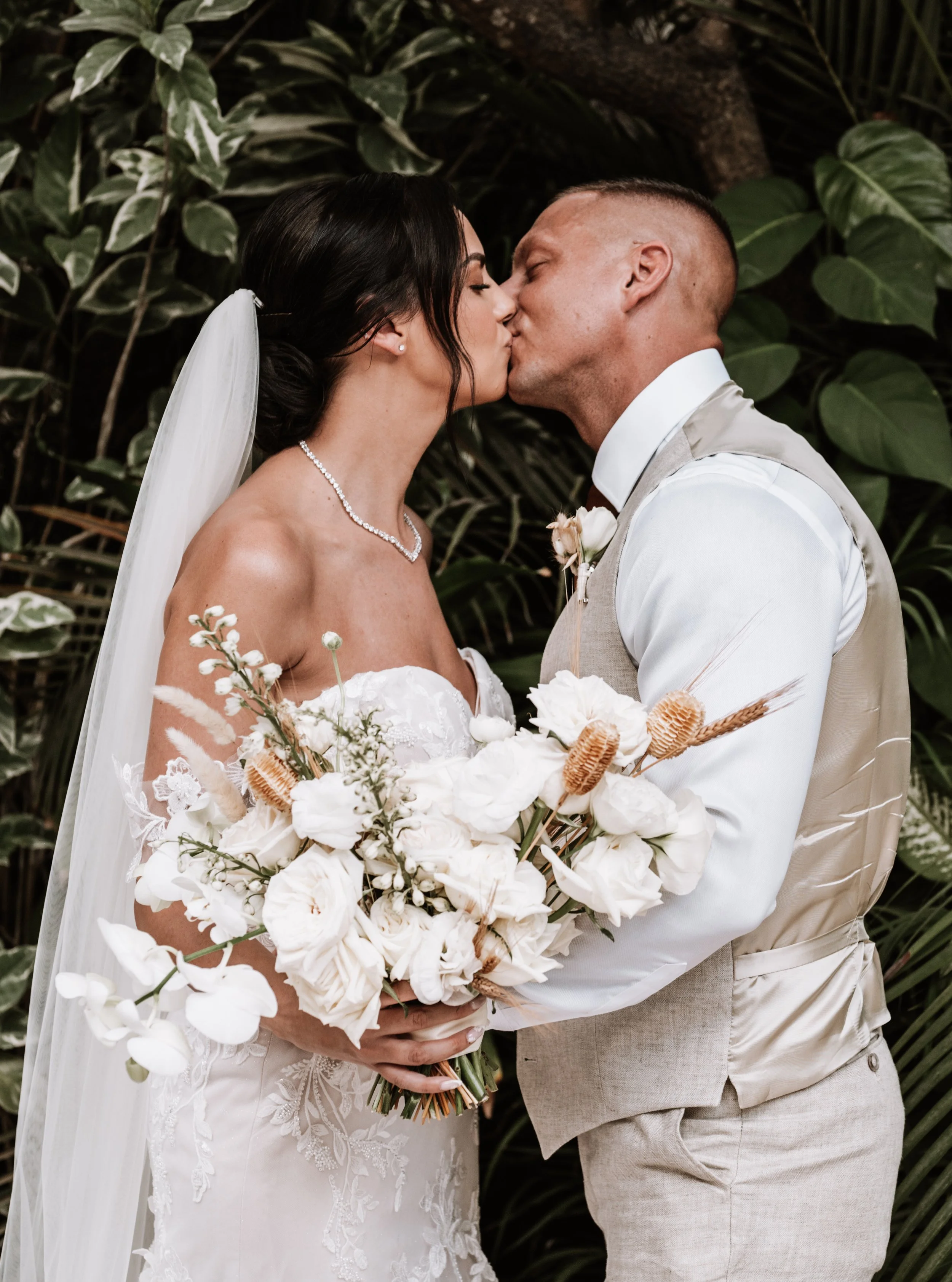 Man and a woman in wedding attire, woman is in a white strapless dress, holding a large bouquet, and man is in a light suit and they are standing close together.