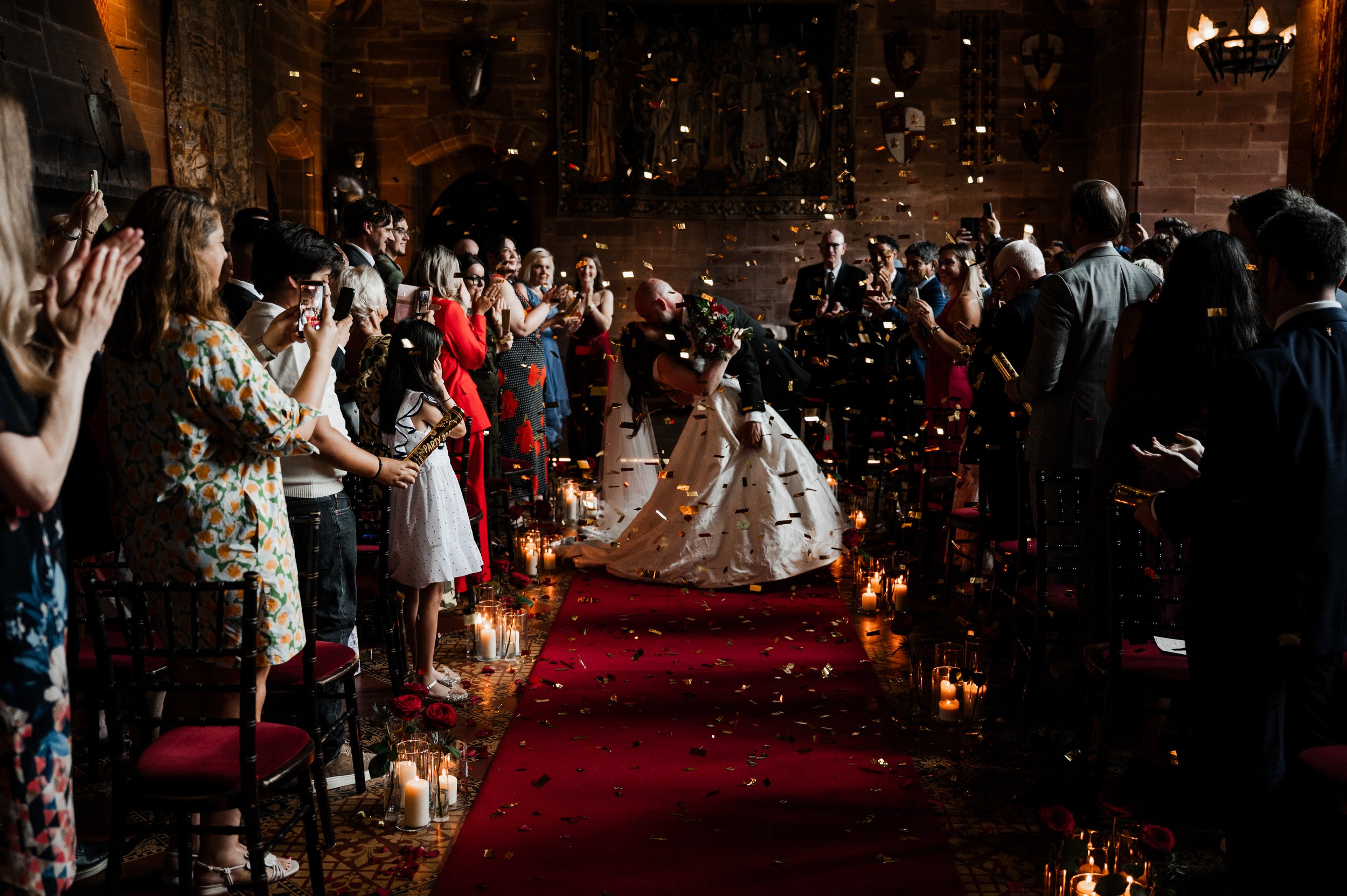 Peckforton Castle -View up an aisle at bride being dipped for a kiss by the groom surrounded by wedding guests in pews with confetti in the air. 