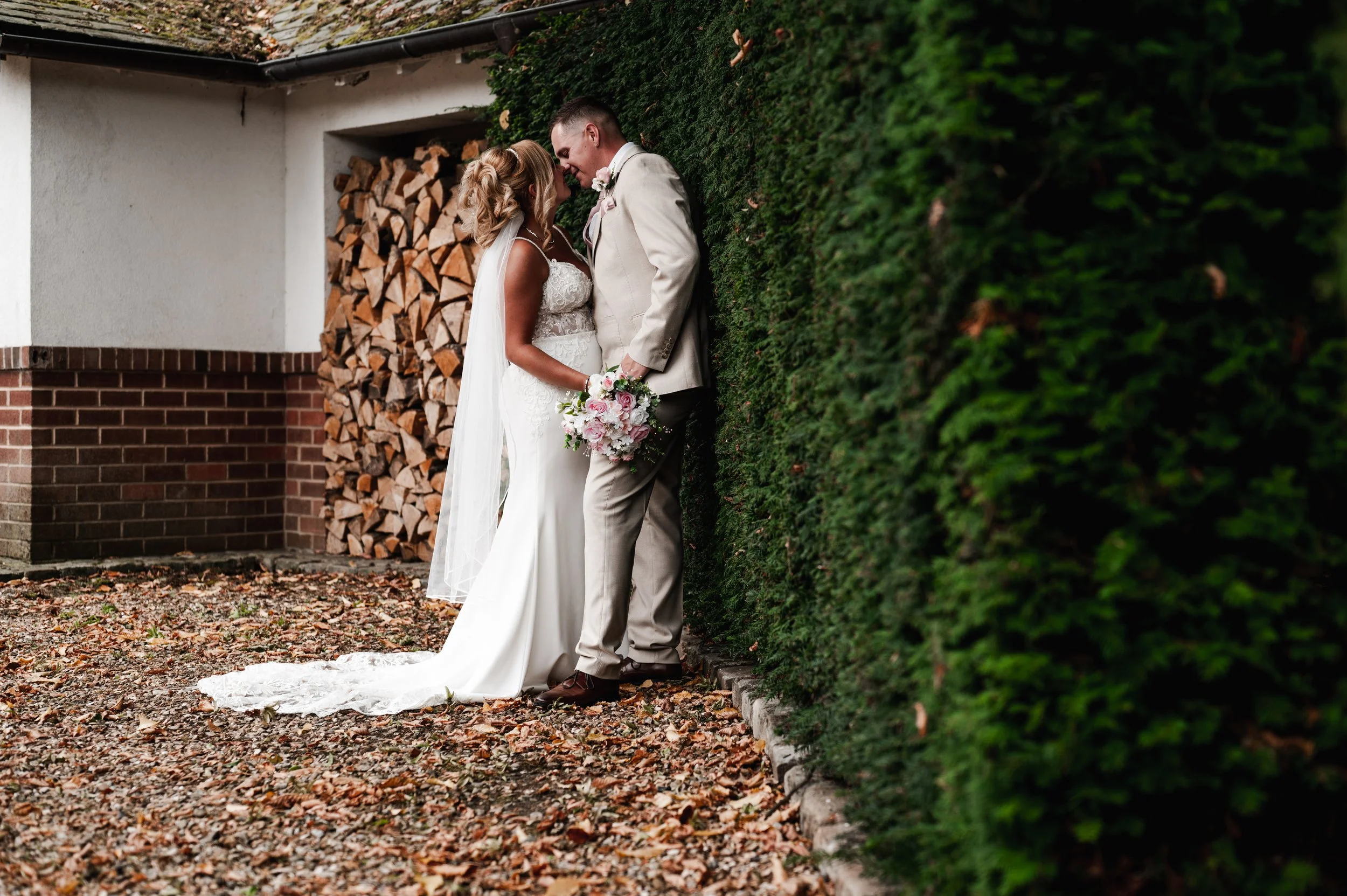 The Love Shack - Groom and bride in wedding attire kissing with a foliage hedge behind them. 
