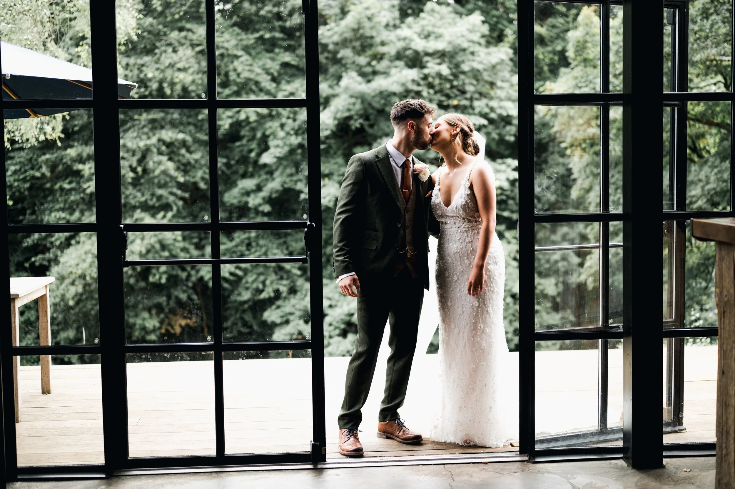 Bride and groom sharing a kiss on a wooden deck viewed through glass doors with greenery in the background hidden river cabins, Gareth roy photography