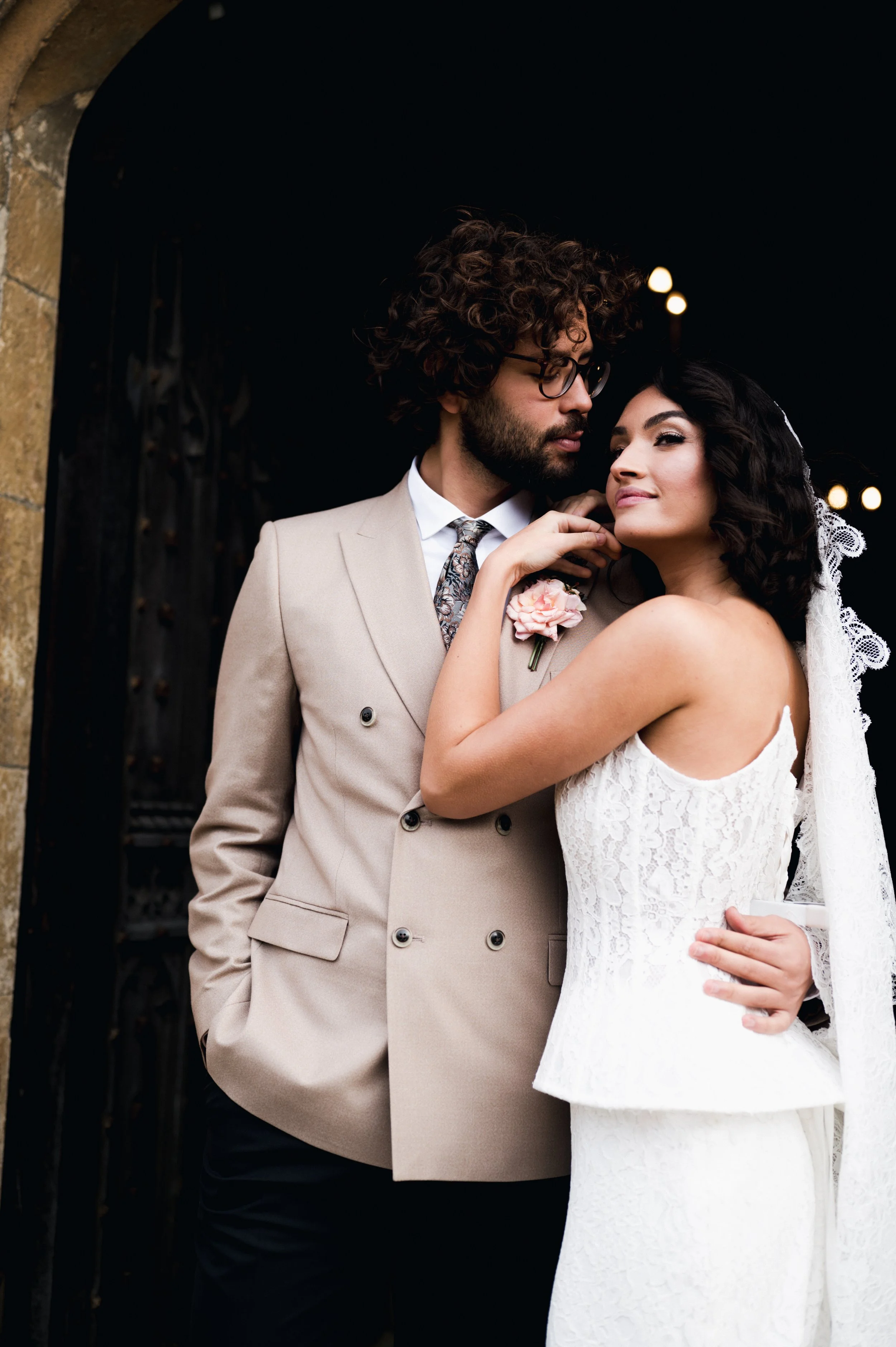 A bride and groom in wedding attire standing close together, with the groom in a beige suit and the bride in a white lace dress and veil, at what appears to be a wedding or special event at Sudeley castle