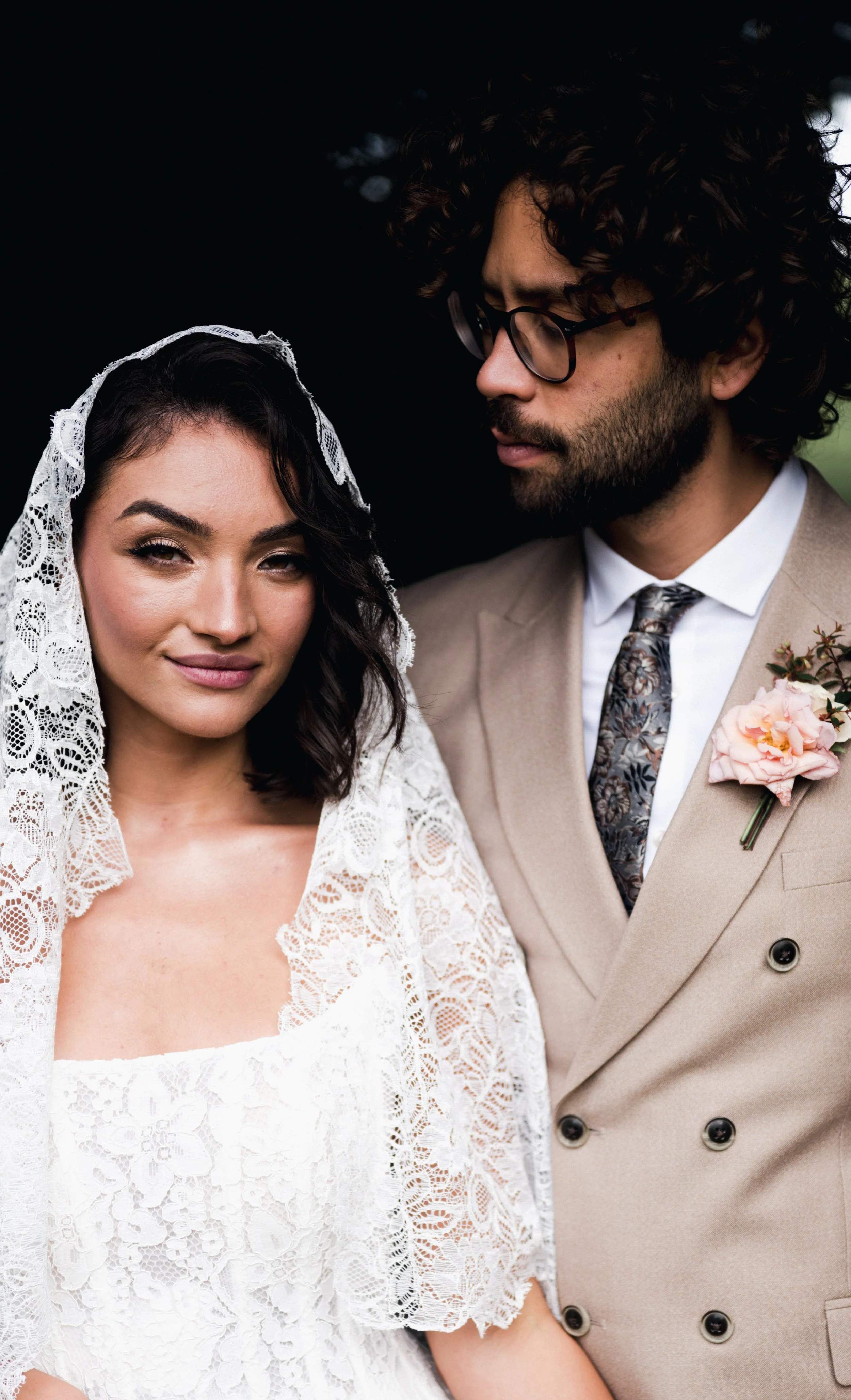 A bride with dark wavy hair wearing a lace wedding dress and veil, and a groom with curly hair, glasses, a beige suit, and a boutonniere, standing close together outdoors at sudeley castle, Gareth roy photography